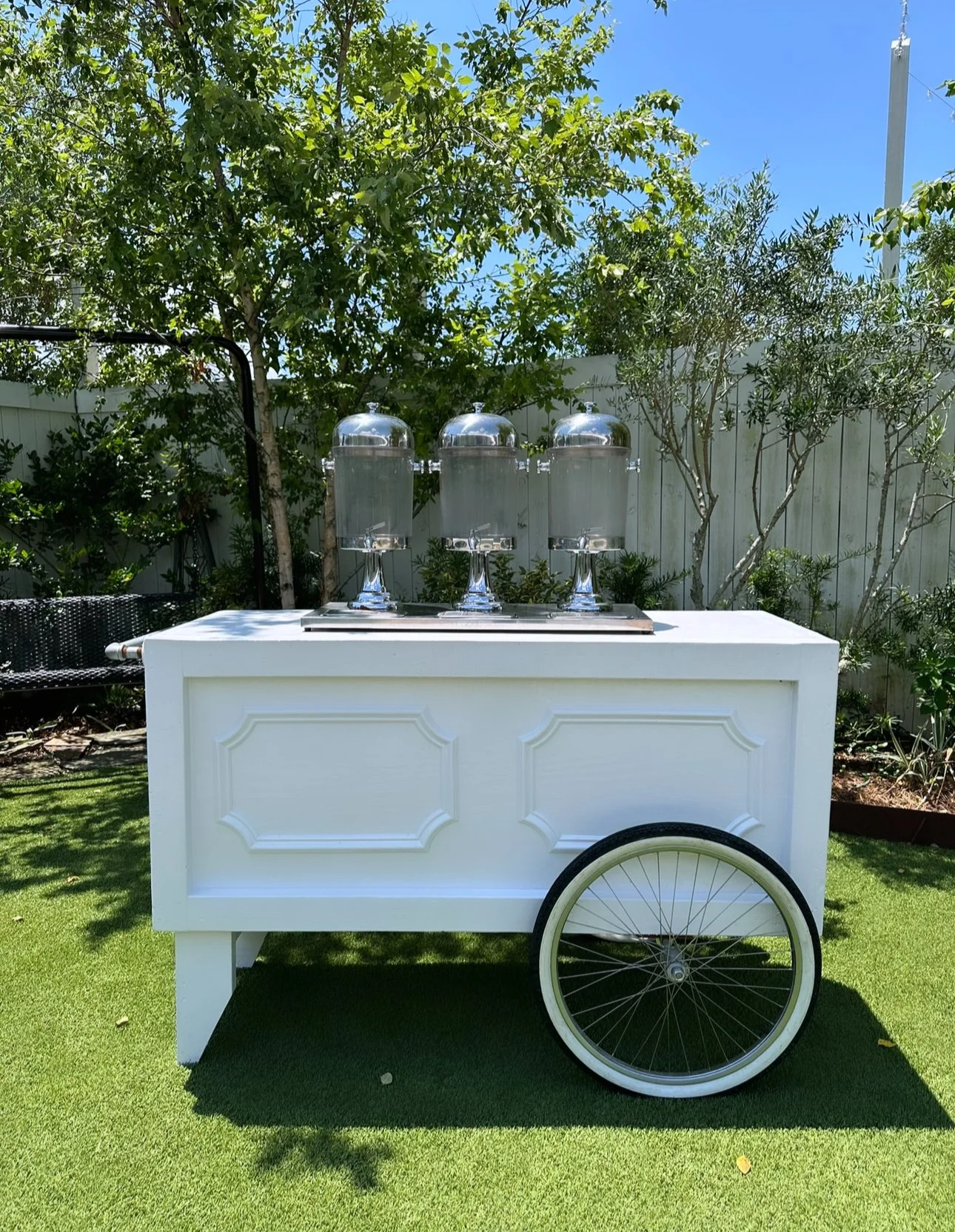 White beverage cart with a large wheel, set outdoors on green artificial grass, with three glass beverage dispensers on top, surrounded by bushes and trees under a blue sky.
