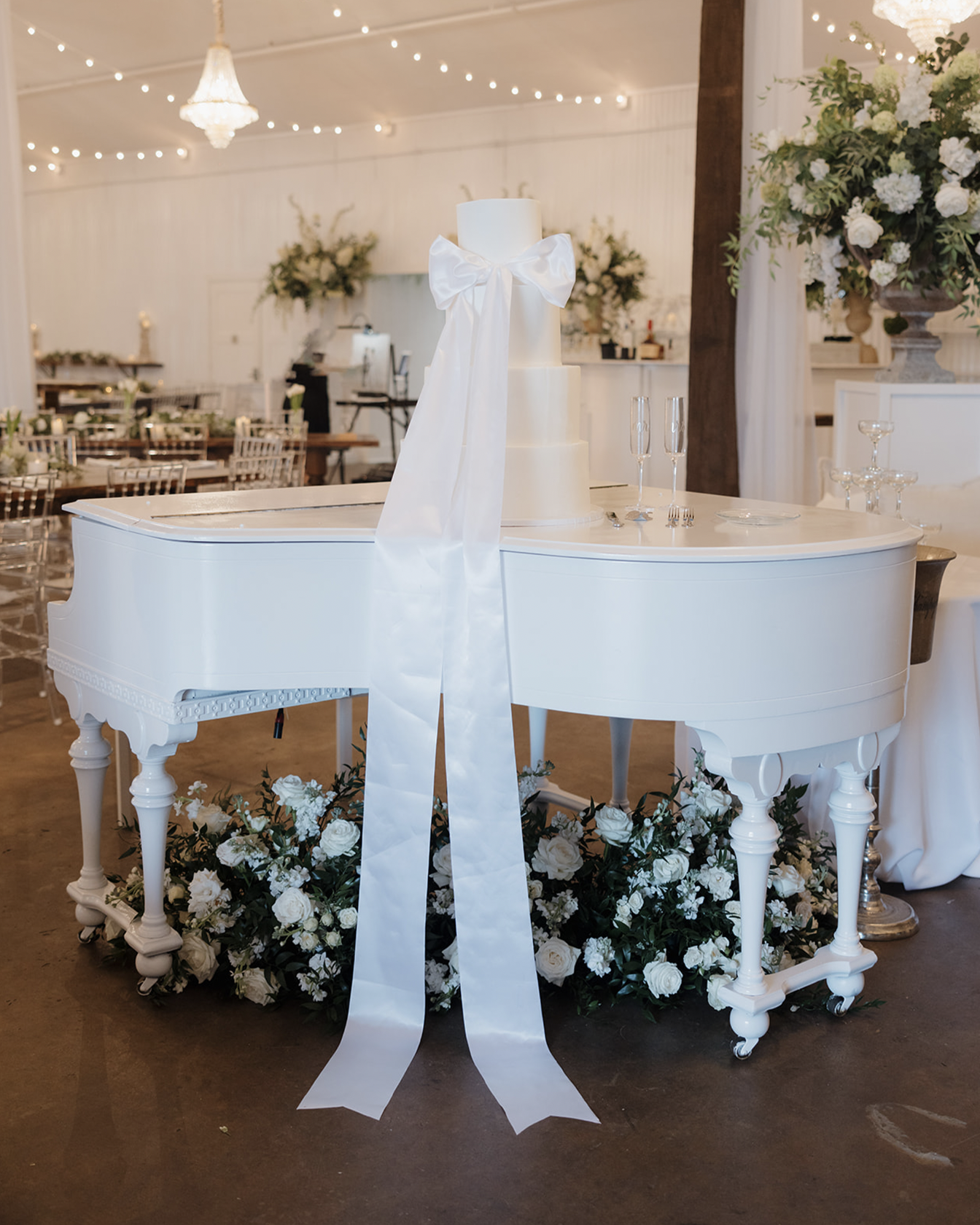 A white wedding cake with three tiers, decorated with a large white satin bow, is positioned on a white grand piano surrounded by white flowers, set in a decorated event hall with string lights and floral arrangements.