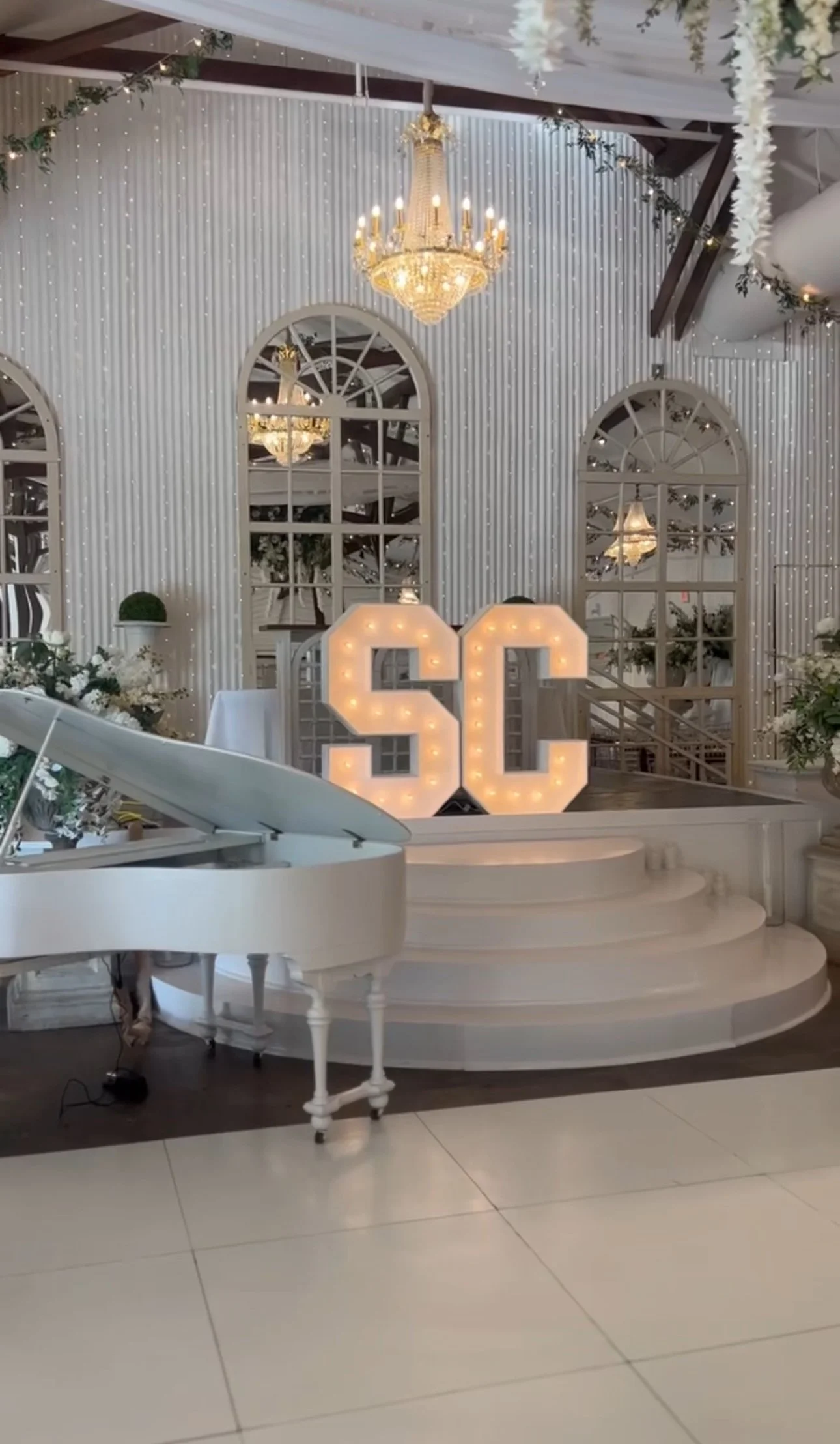 Decorative indoor space with a white grand piano, illuminated sign with the letters 'SC' on a round stage, large ornate chandelier hanging from the ceiling, large arched windows, and Christmas garland decorations.