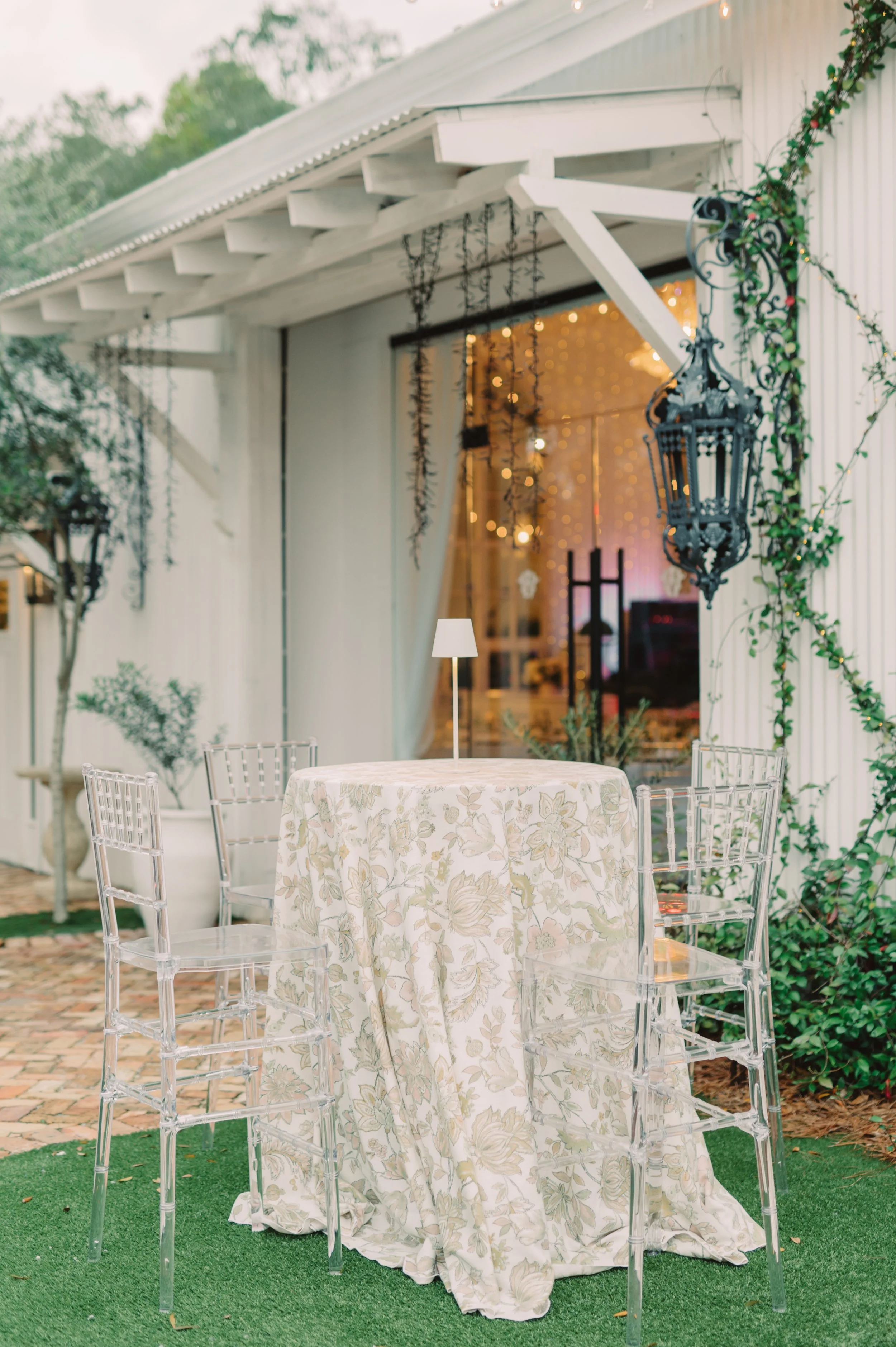 An outdoor patio in a Pensacola Wedding Venue area with a round table covered in a floral tablecloth, surrounded by clear acrylic chairs. There is a small table lamp on the table and decorative hanging lanterns and plants.