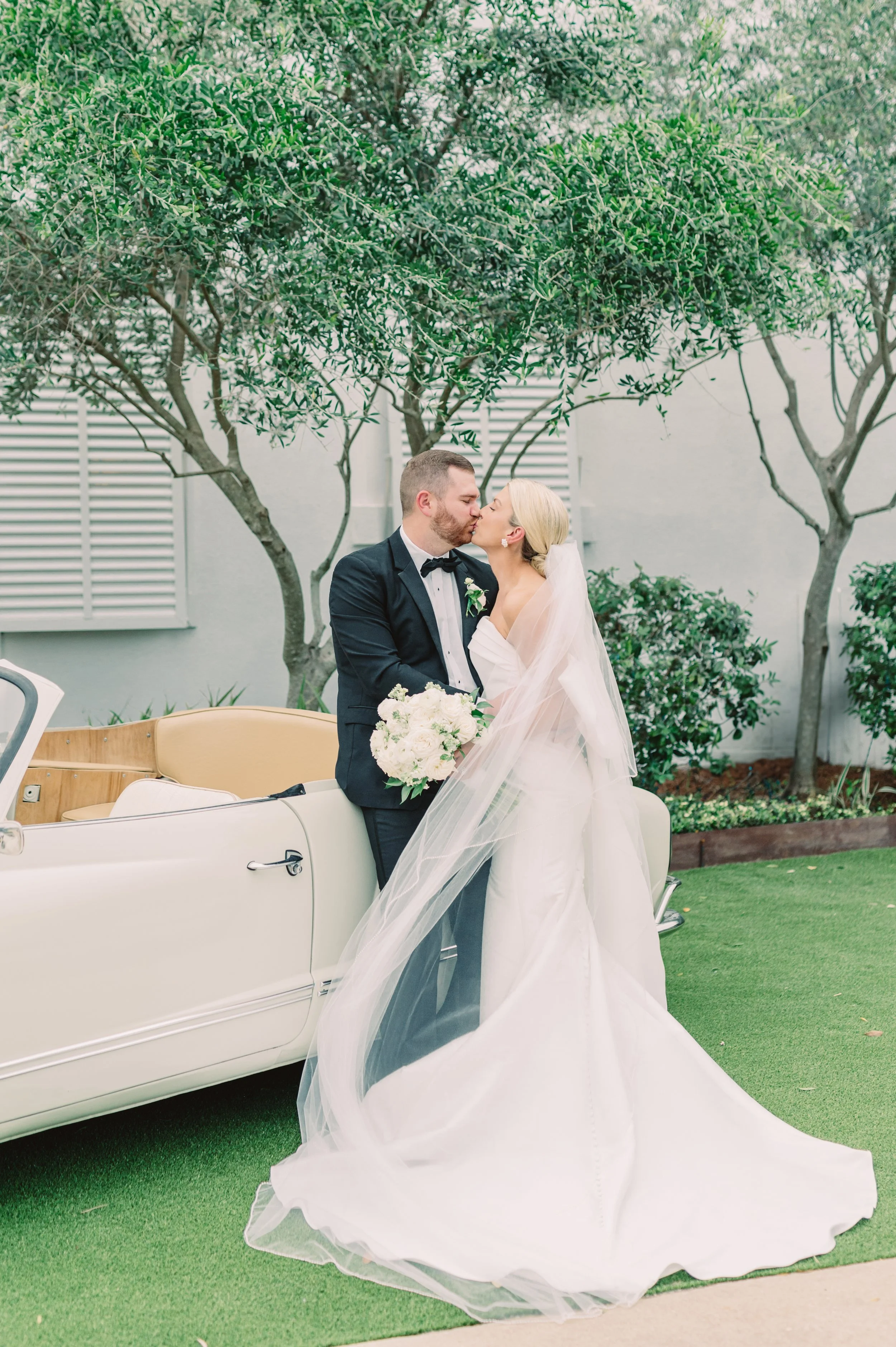 Bride and groom sharing a kiss next to a vintage white car on a lush green lawn with trees in the background of a Pensacola Wedding Venue.