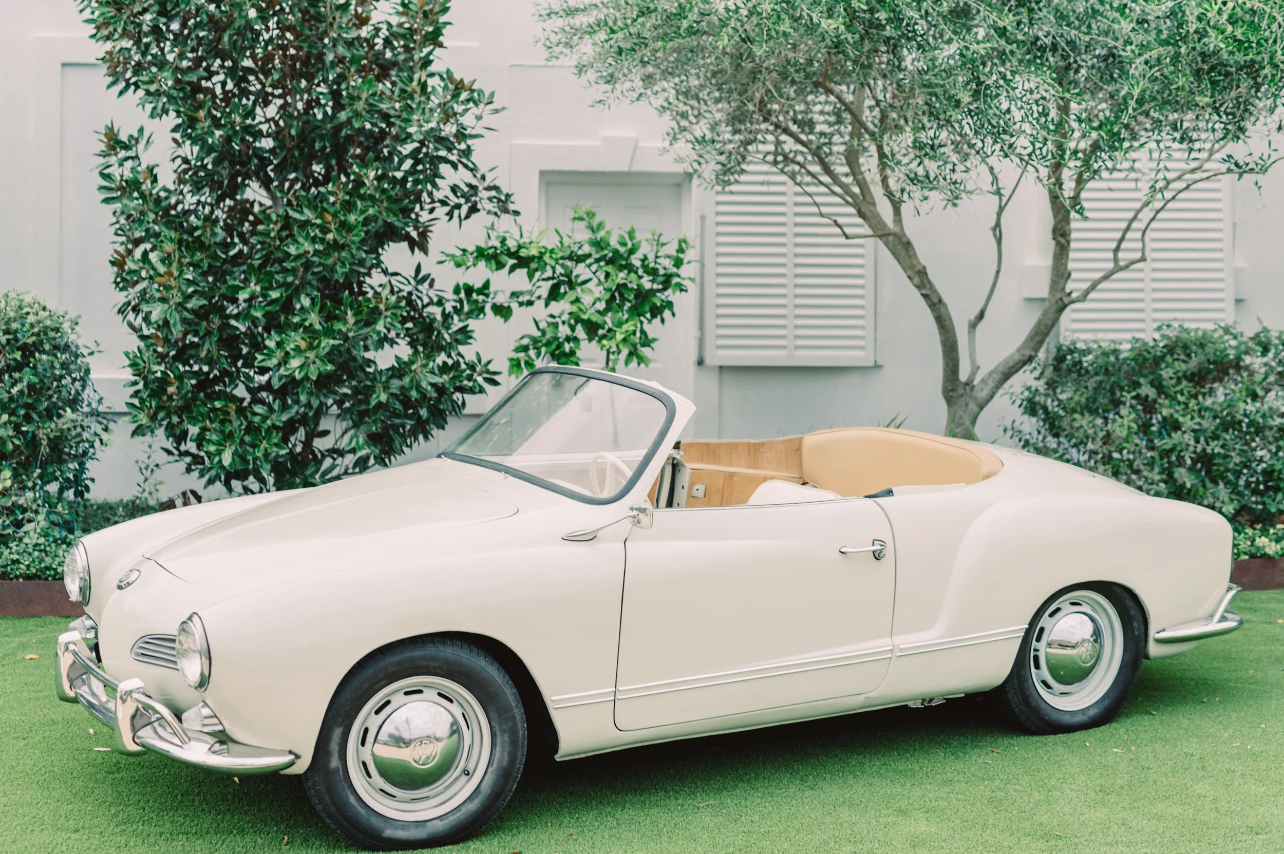 A vintage white convertible car parked on green grass, with trees and a house with white shutters in the background.