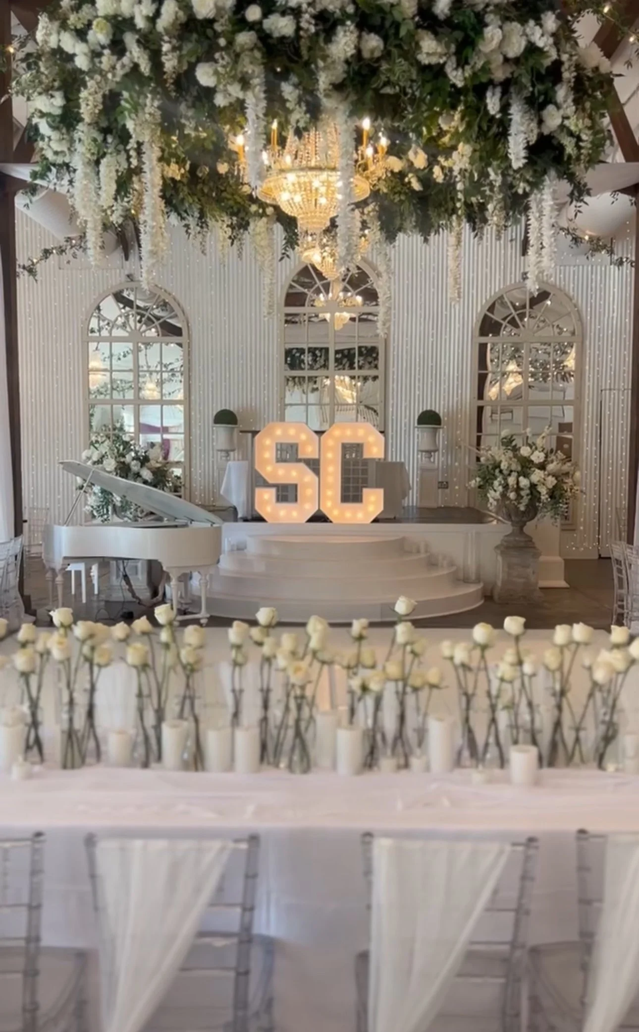 Elegant indoor event space decorated with white flowers and candles, featuring large illuminated initials 'SC' on stage, a white grand piano, and a chandelier, indicating celebratory atmosphere in a Pensacola Wedding Venue