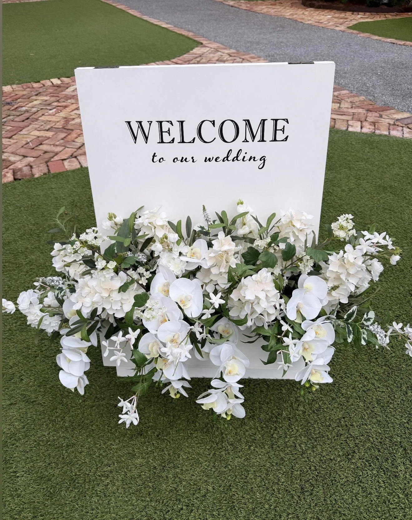 White floral arrangement with a welcome sign for a wedding on green grass with brick patio and paved path in the background.