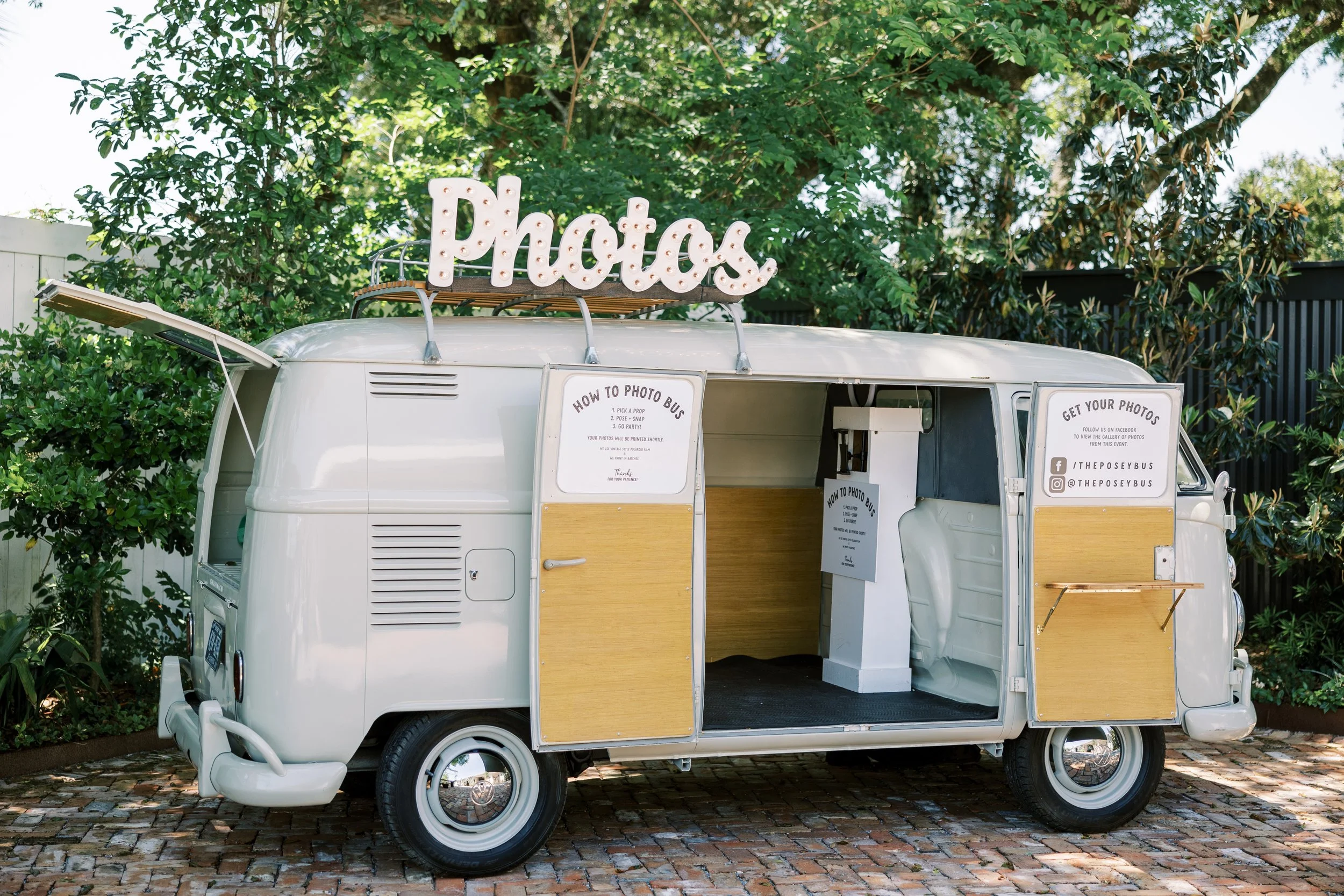 A vintage white Volkswagen van with yellow panels on the sides, converted into a photo booth. The van is parked on a brick-paved area surrounded by trees and greenery. There is a lit-up sign on top spelling out "Photos". Open doors reveal the interio