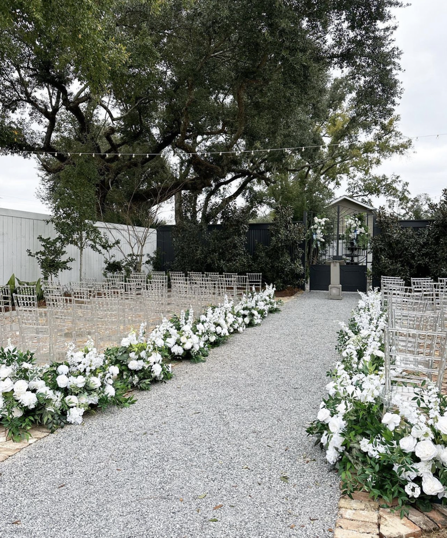 Outdoor wedding setup with white flowers along the aisle, clear chairs, and a black and white altar at the end of the gravel pathway, surrounded by trees and greenery.