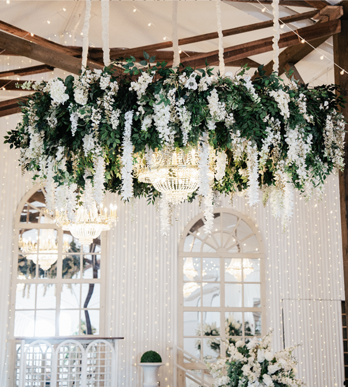 Elegant chandelier hanging amidst a lush arrangement of white flowers and greenery, with a backdrop of warm string lights and arched windows.