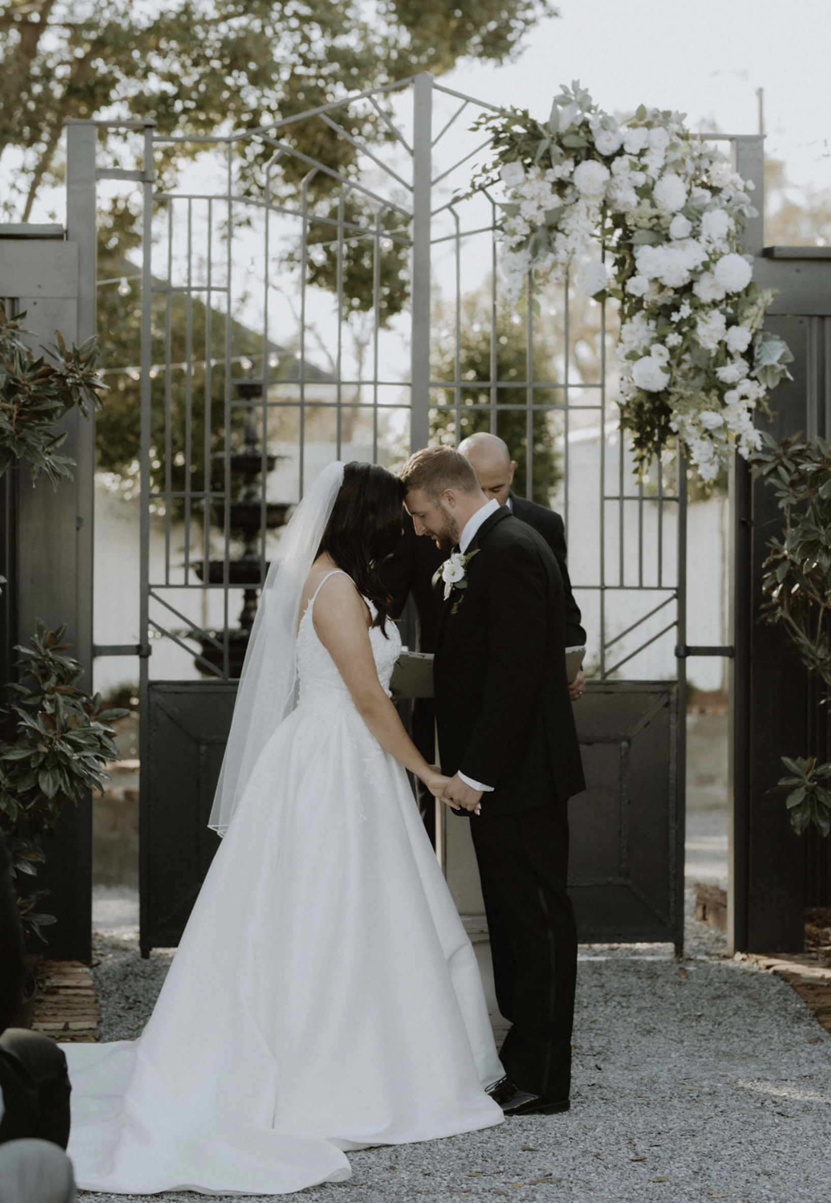 A wedding ceremony with a bride and groom holding hands, bowing their heads, and standing before an officiant outdoors with a metal gate decorated with white flowers in the background.
