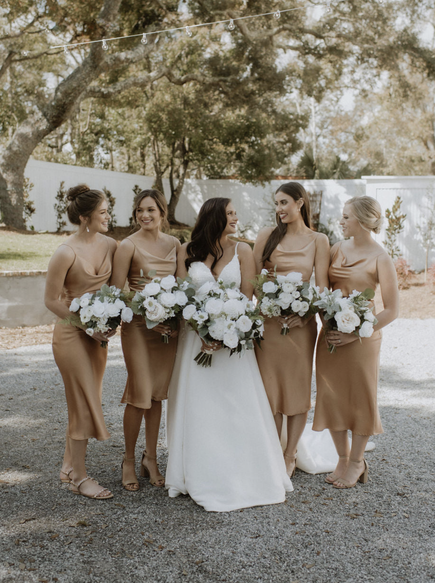 A bride and five bridesmaids standing outdoors in a garden area, all holding bouquets of white flowers, and smiling at each other.