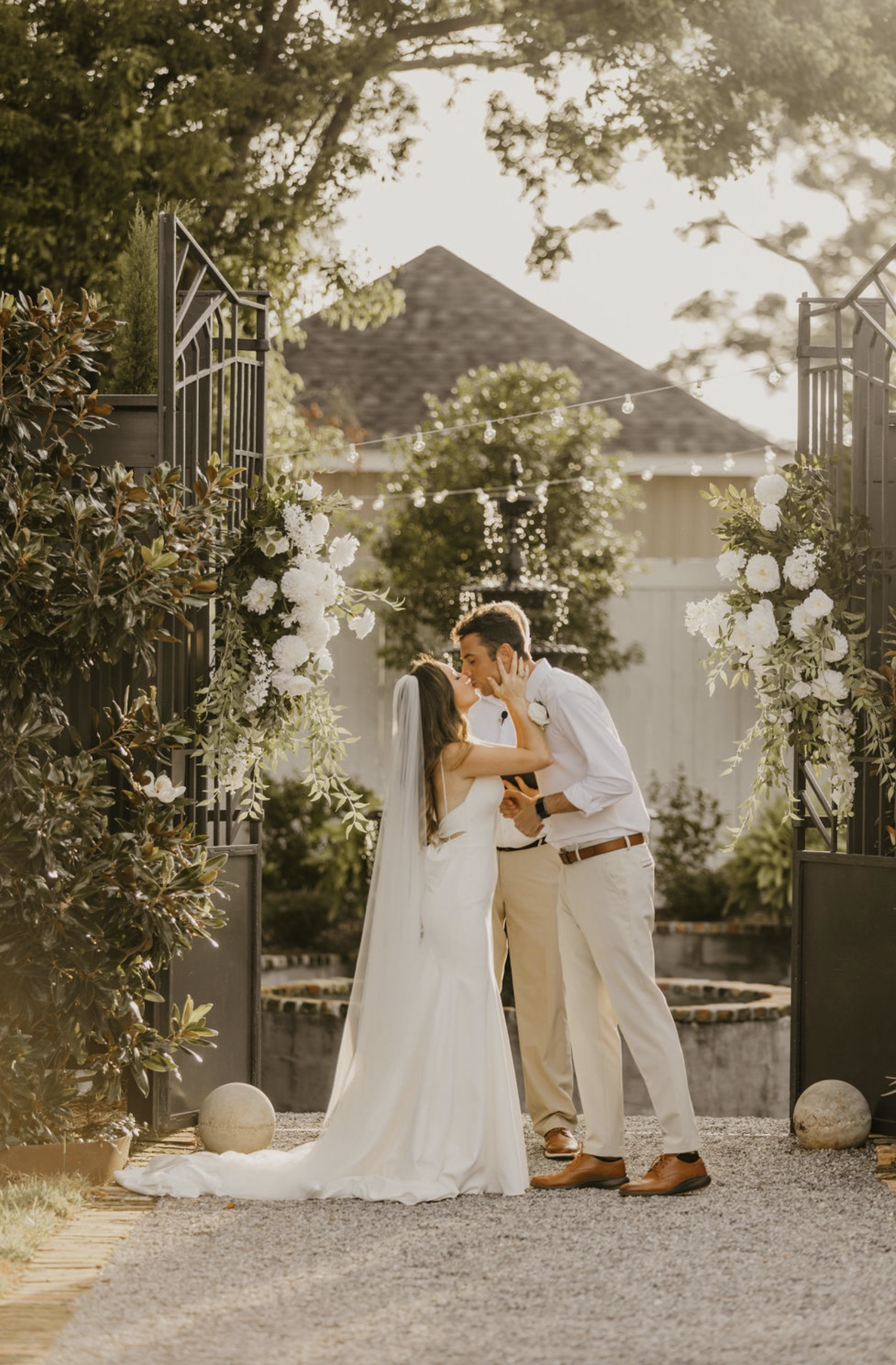 A bride and groom kissing during their wedding ceremony outdoors, framed by a black metal gate decorated with white flowers, with greenery and a fountain in the background.