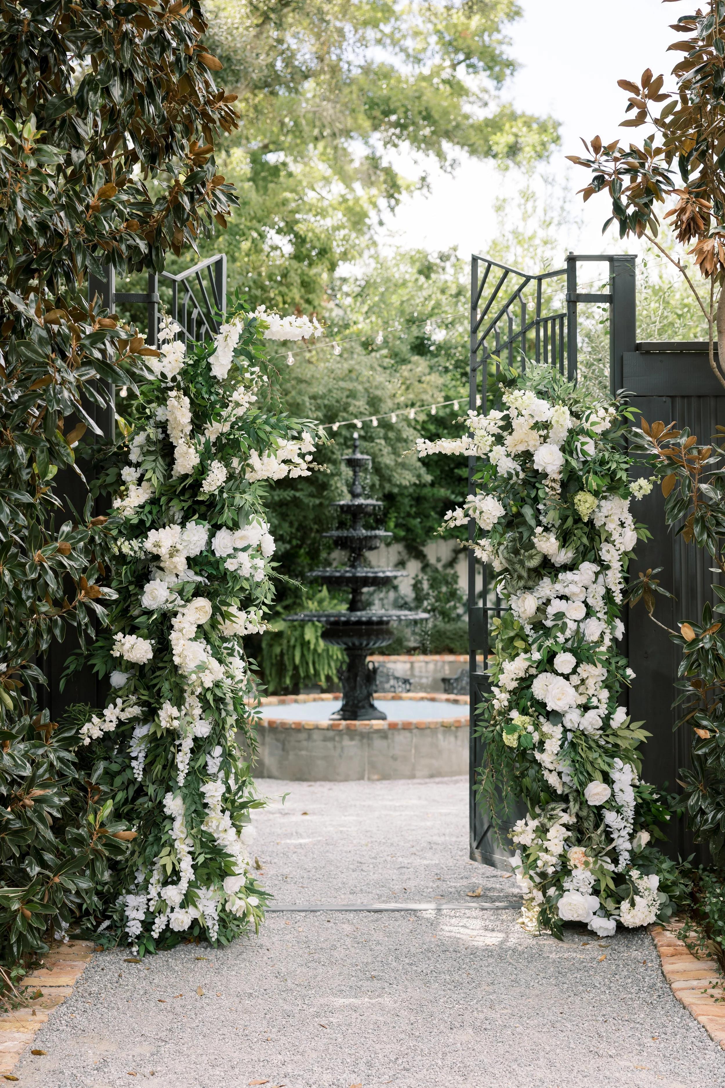 Decorated entrance with black metal gates adorned with white flowers and greenery, leading to a fountain in a lush garden.