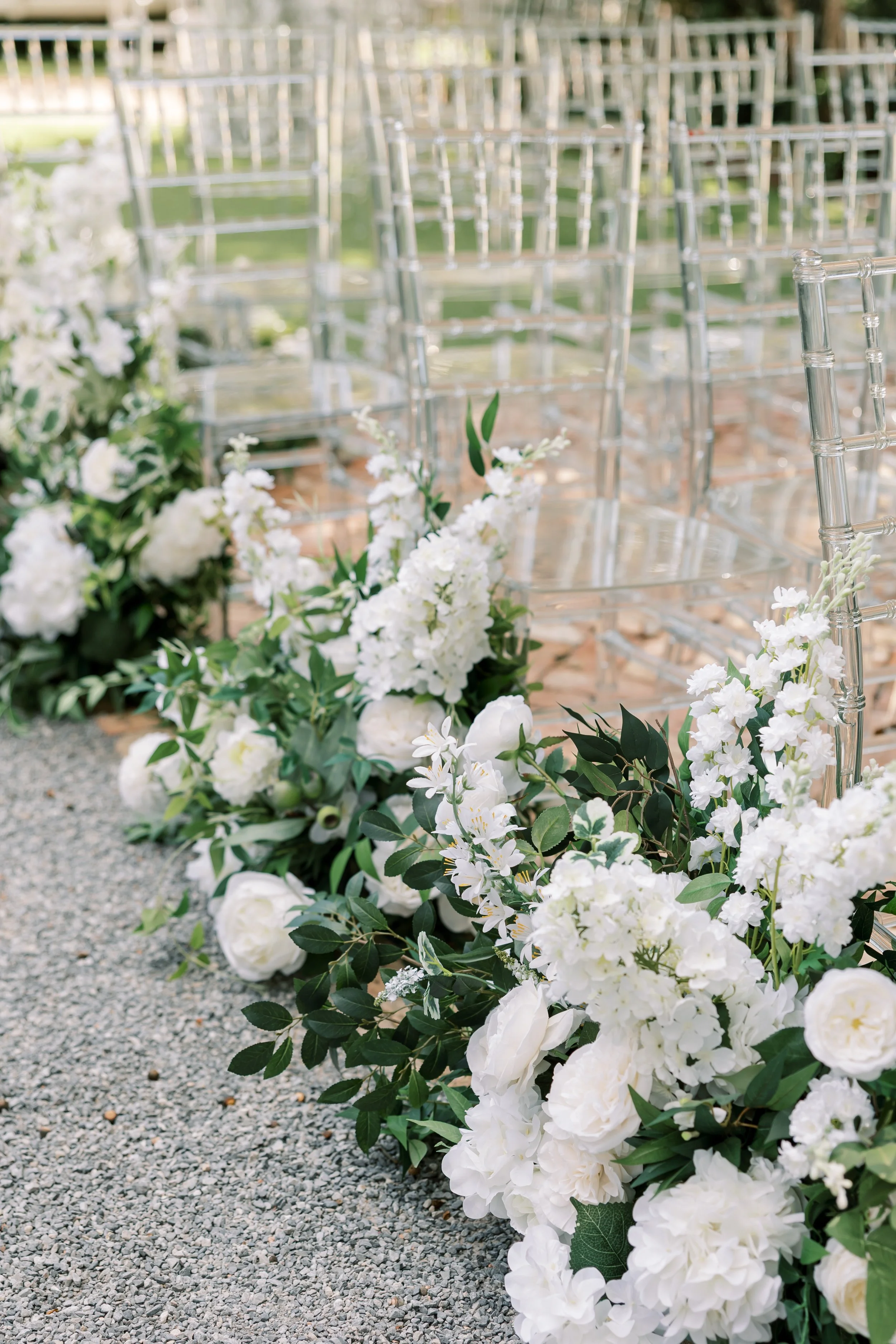 White floral arrangements along a gravel aisle with transparent chairs in the background, set for a wedding ceremony in Pensacola.