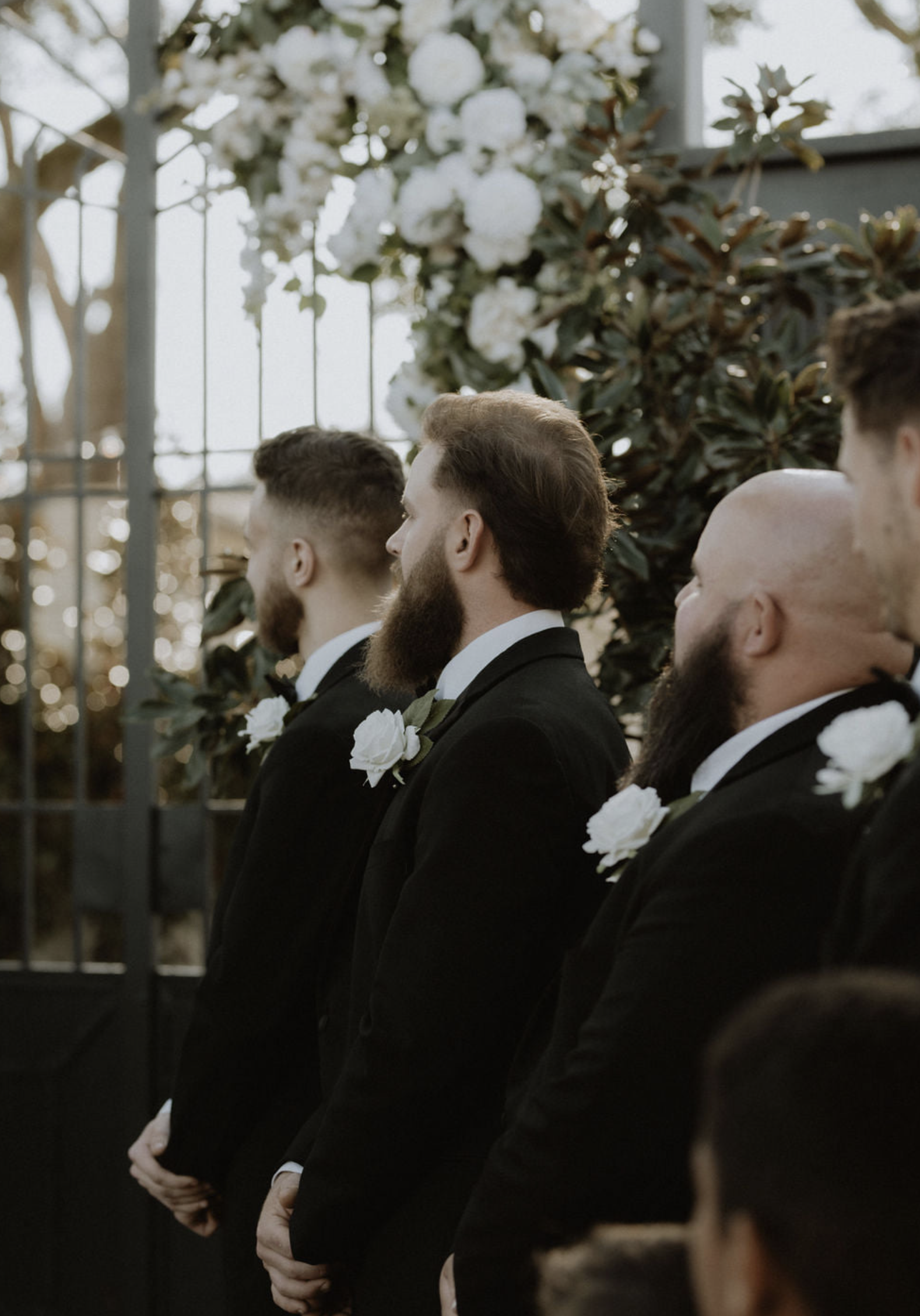 Groomsmen in black tuxedos with white boutonnieres standing in a line during a wedding ceremony outdoors, with greenery and floral arrangements in the background.