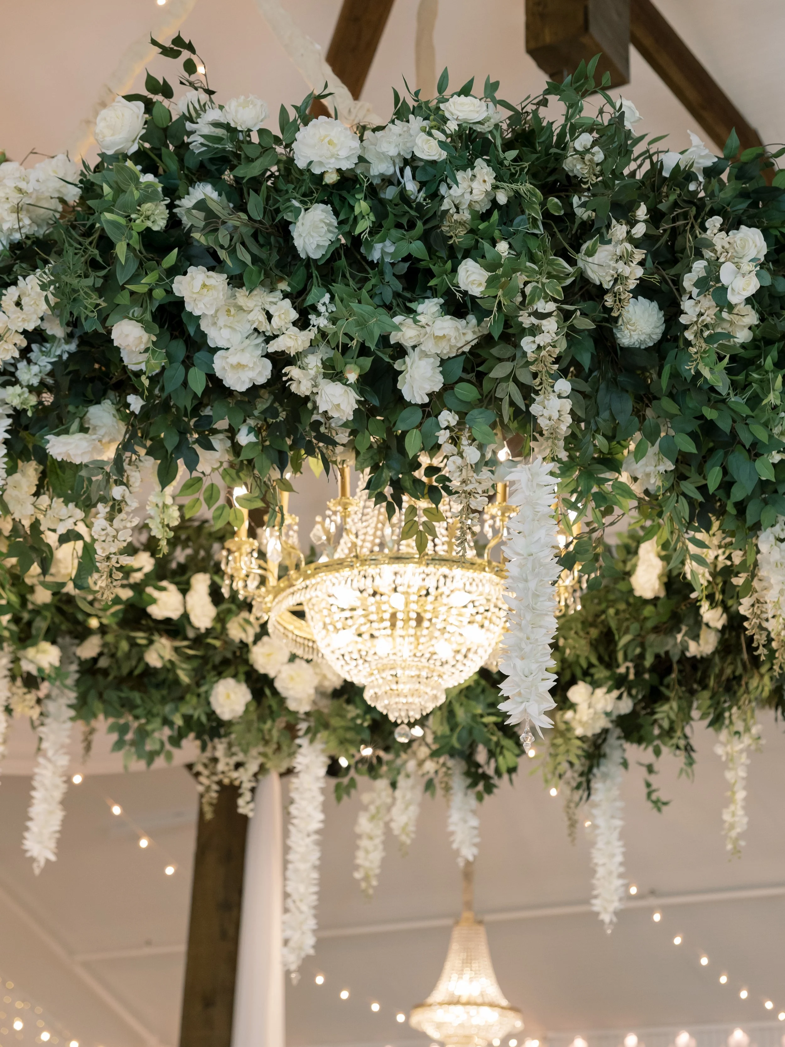 Elegant floral chandelier with white flowers and greenery hanging from ceiling, with two chandeliers below and string lights in the background.
