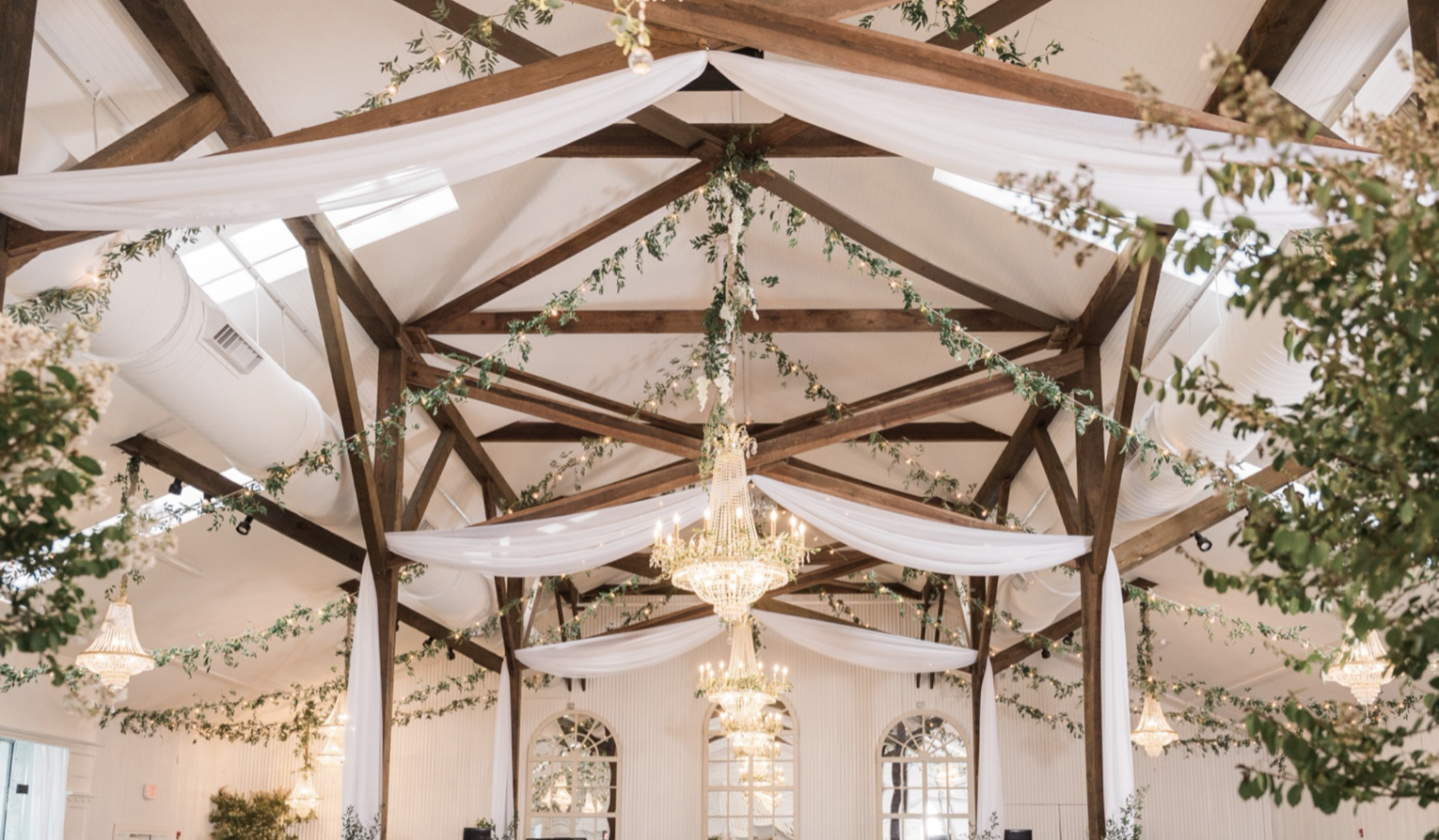 Decorated wedding banquet hall with chandeliers, draped white fabric, greenery garlands hanging from a wooden ceiling structure, and large arched windows.