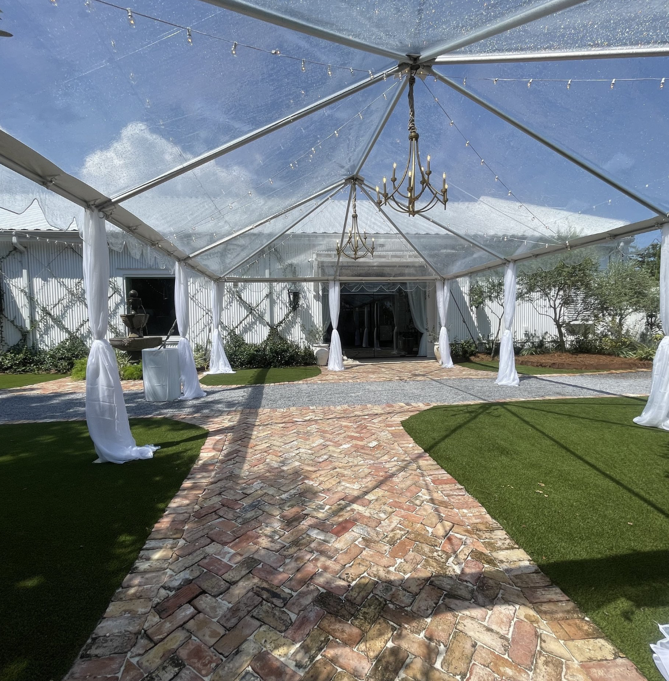An outdoor event space with a transparent tent, brick pathway, greenery, chandeliers, and string lights.