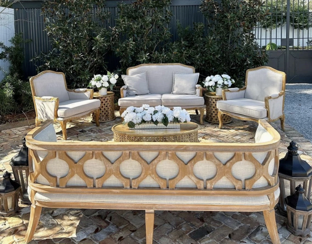 Outdoor seating area with a cream and gold vintage furniture set, including a curved sofa, two armchairs, and a coffee table with white floral arrangements, on a brick patio surrounded by shrubs.