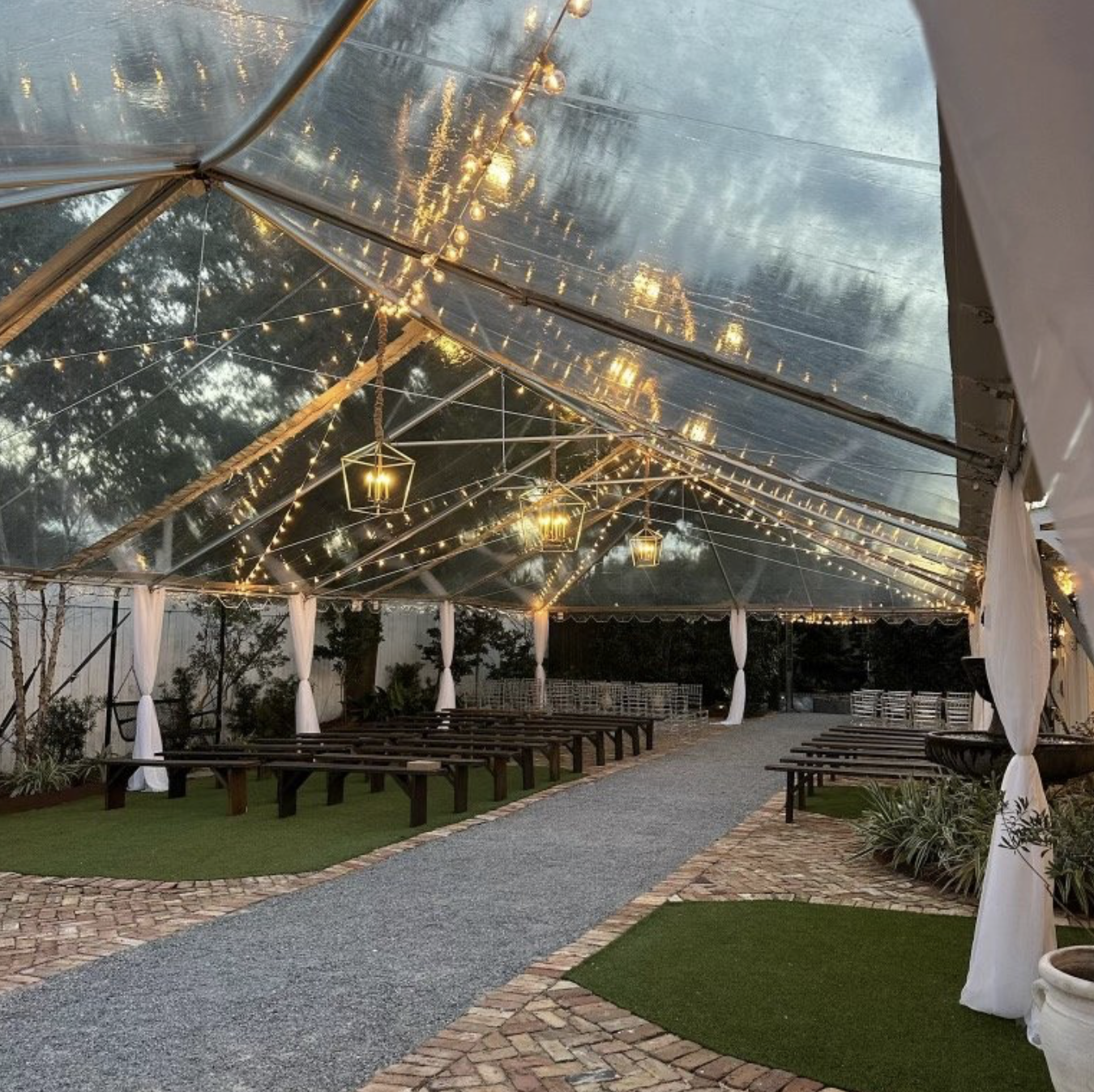 Indoor event tent decorated with string lights and hanging lanterns, with wooden benches, greenery, and a paved pathway in a Pensacola Wedding Venue.
