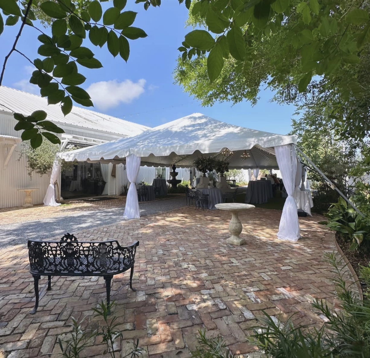 Outdoor celebration area with a large white tent, decorated tables, a black ornate bench, and a birdbath on a brick patio, surrounded by greenery and trees under a blue sky.