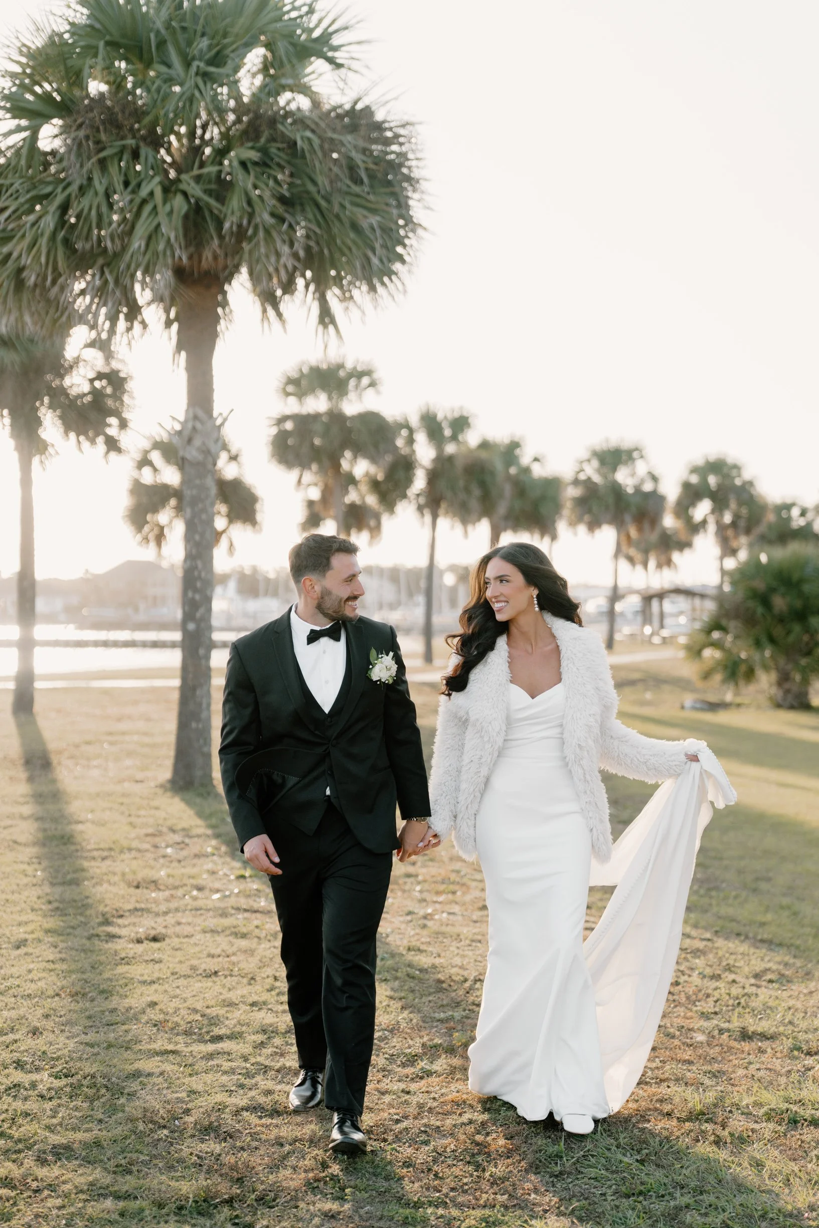 A Pensacola bride and groom walking hand in hand outdoors near palm trees at sunset, smiling at each other. The bride is wearing a white wedding dress with a fur coat, and the groom is in a black tuxedo with a bow tie.