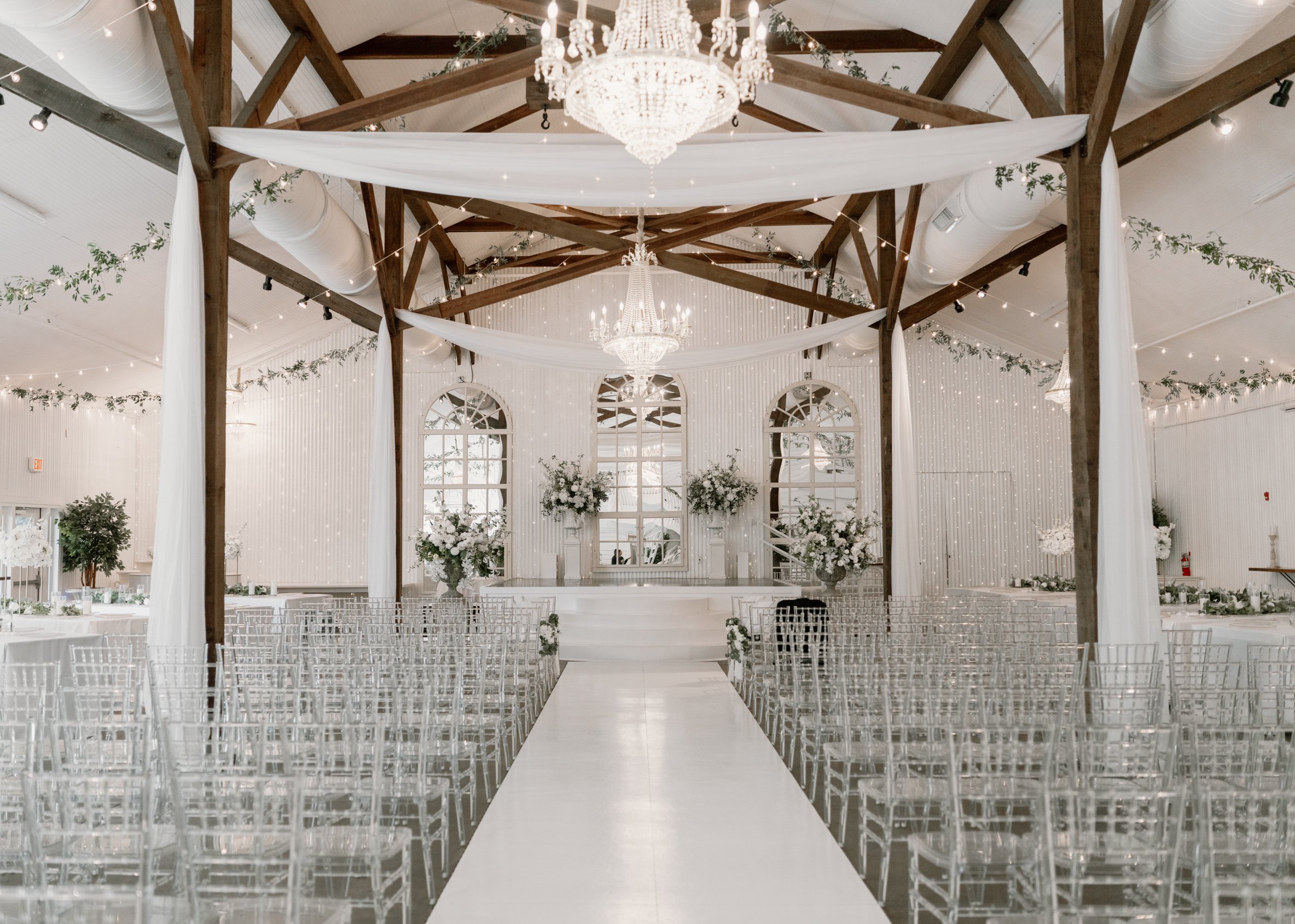 Elegant wedding ceremony setup in a bright, white venue with wooden beams, chandeliers, floral arrangements, and rows of transparent chairs in a Pensacola Wedding Venue.