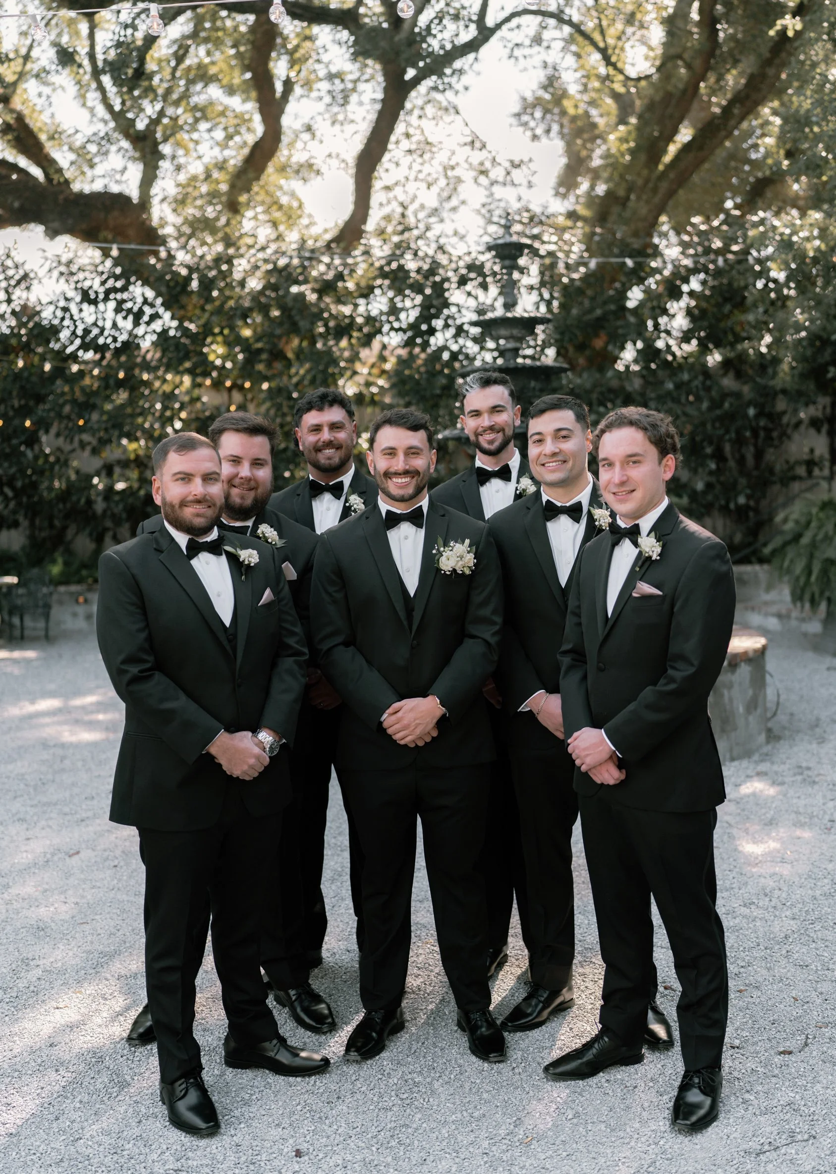 Group of seven men dressed in black tuxedos with bow ties, standing outdoors in a garden setting with trees and a fountain in the background, smiling at the camera in a Pensacola Wedding Venue.