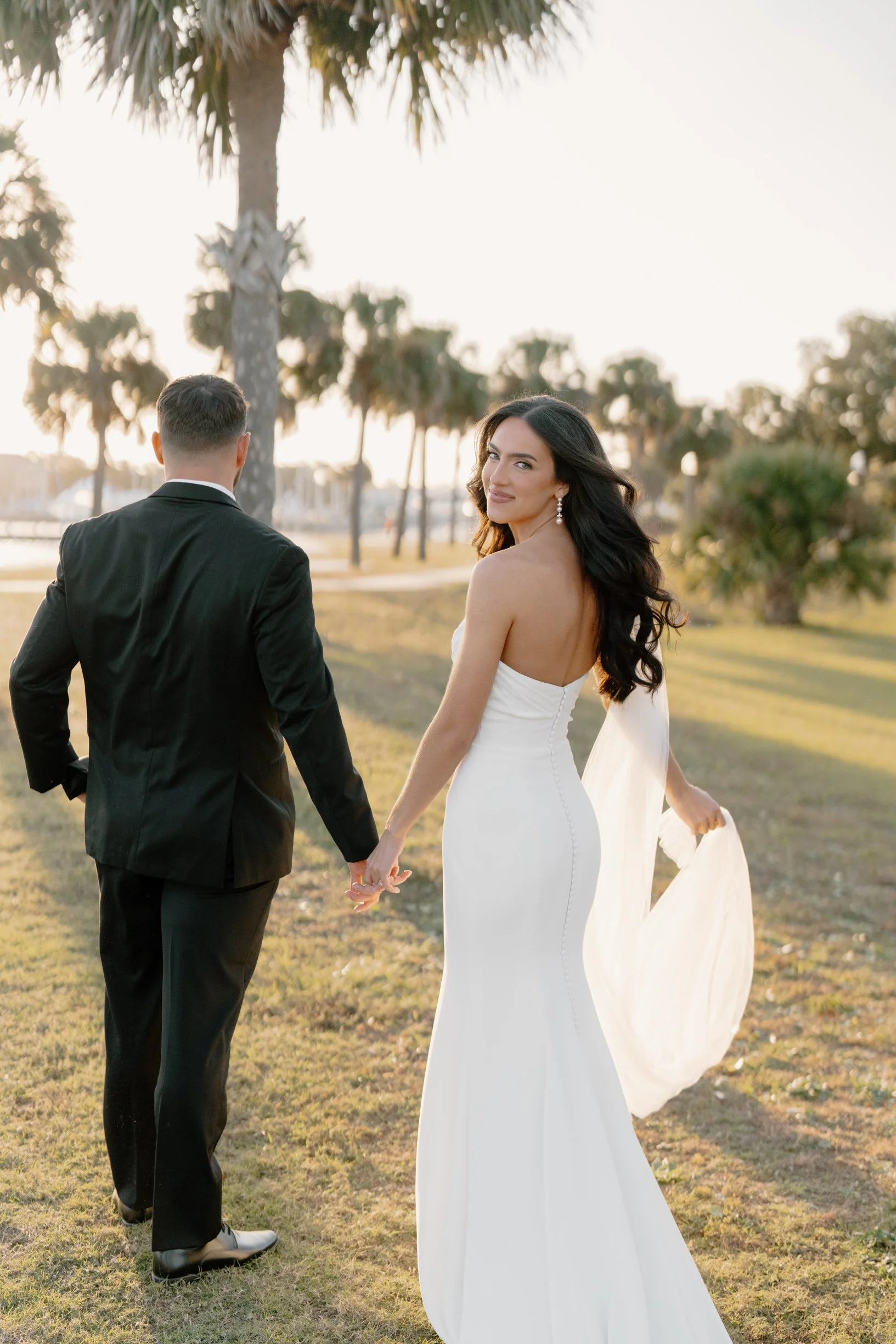 Bride in a white wedding gown holding hands with groom in a black tuxedo, walking outdoors on grass with palm trees and a sunset in the background.