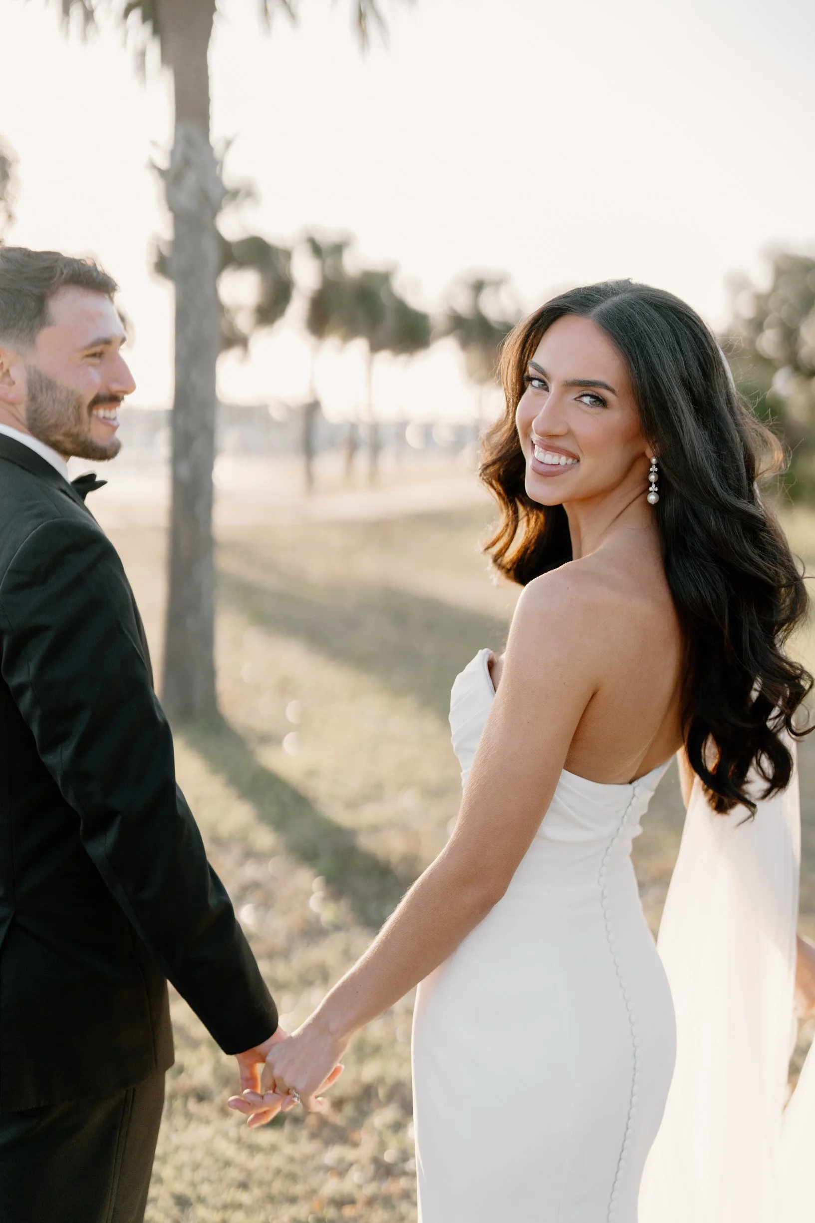 A bride and groom holding hands outdoors during sunset, with trees and a blurred background, smiling at each other.