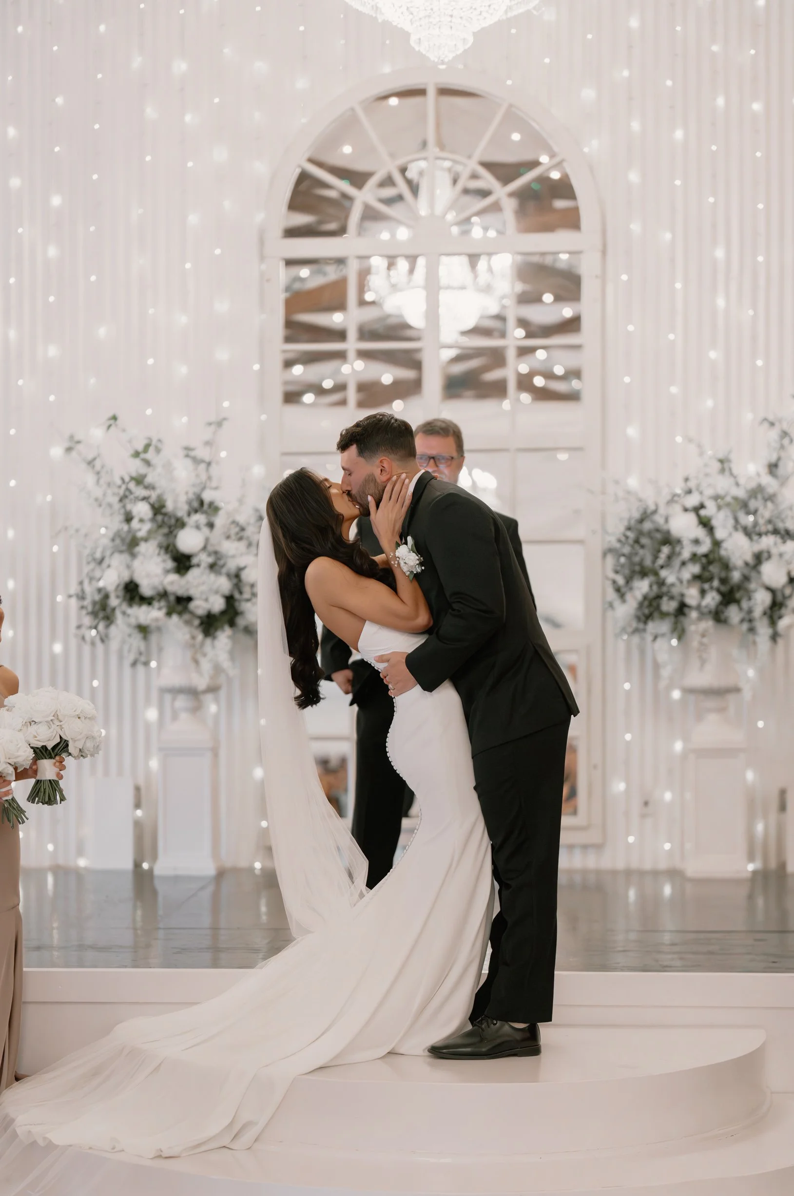 A bride and groom share a kiss during their wedding ceremony in a decorated indoor venue, with floral arrangements and a large mirror behind them.