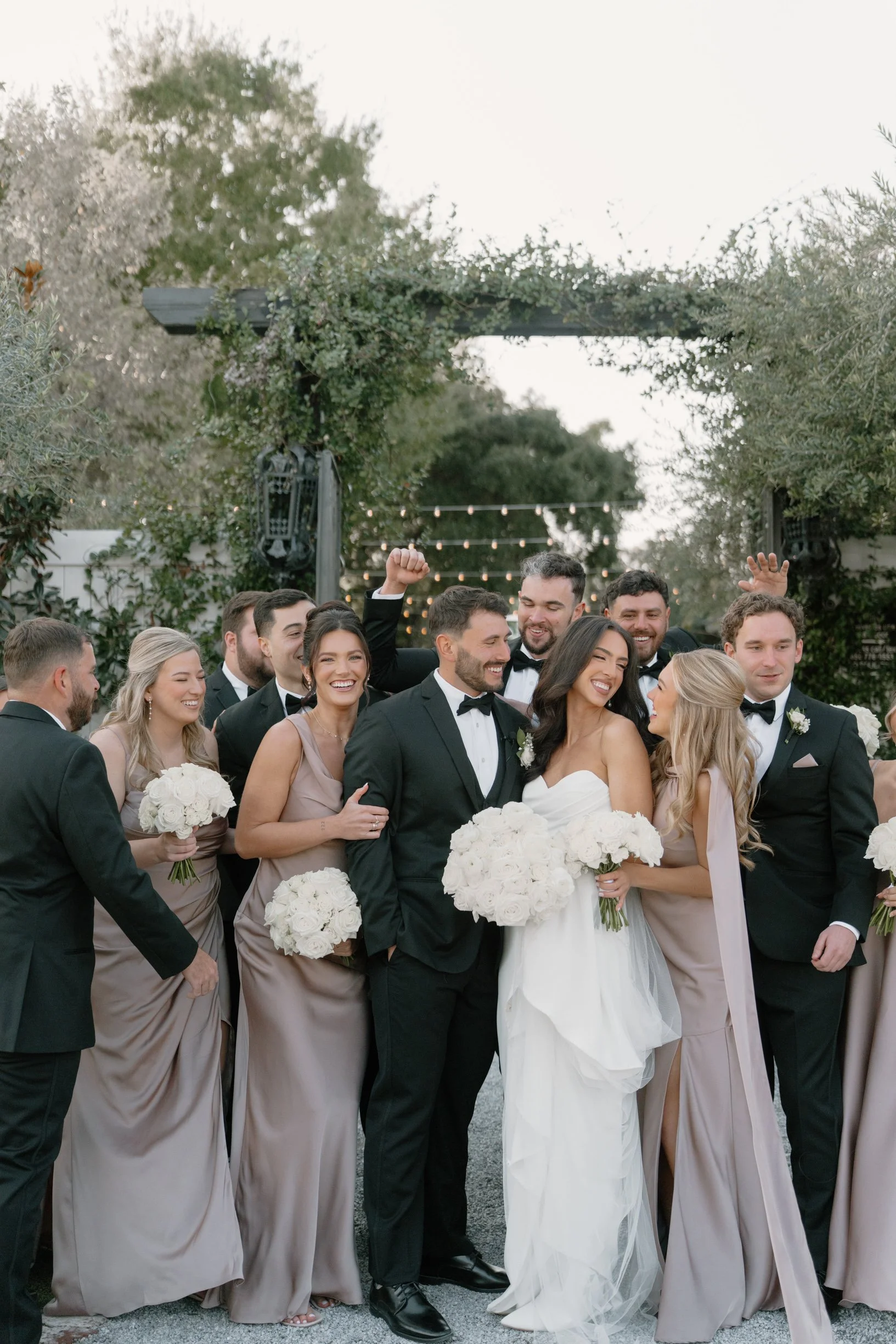 Group of wedding guests dressed in formal attire, celebrating outdoors with smiling faces, at a wedding reception or celebration with fairy lights and trees in the background.