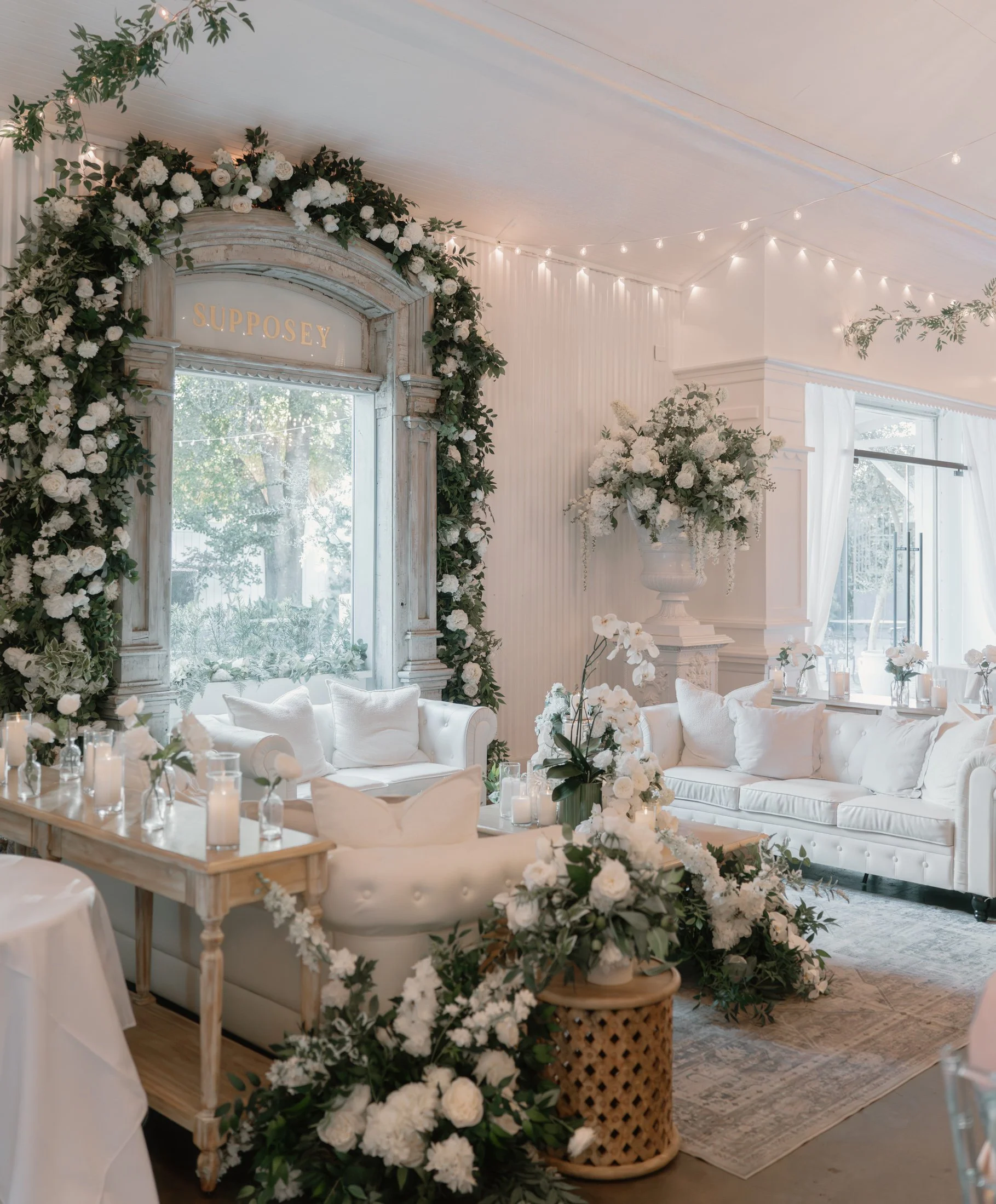 Elegant wedding or event reception with white sofas, floral arrangements, candles, and string lights inside a decorated room with large window and greenery outside.