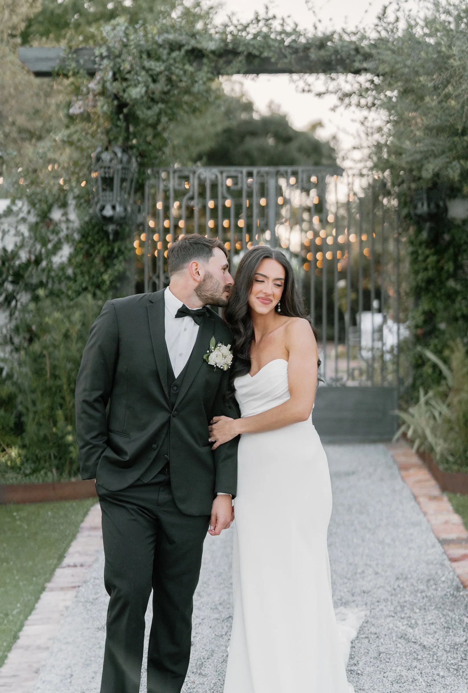 A bride and groom standing on a gravel pathway in a garden, with string lights and greenery in the background, sharing an intimate moment in a Pensacola Wedding Venue.