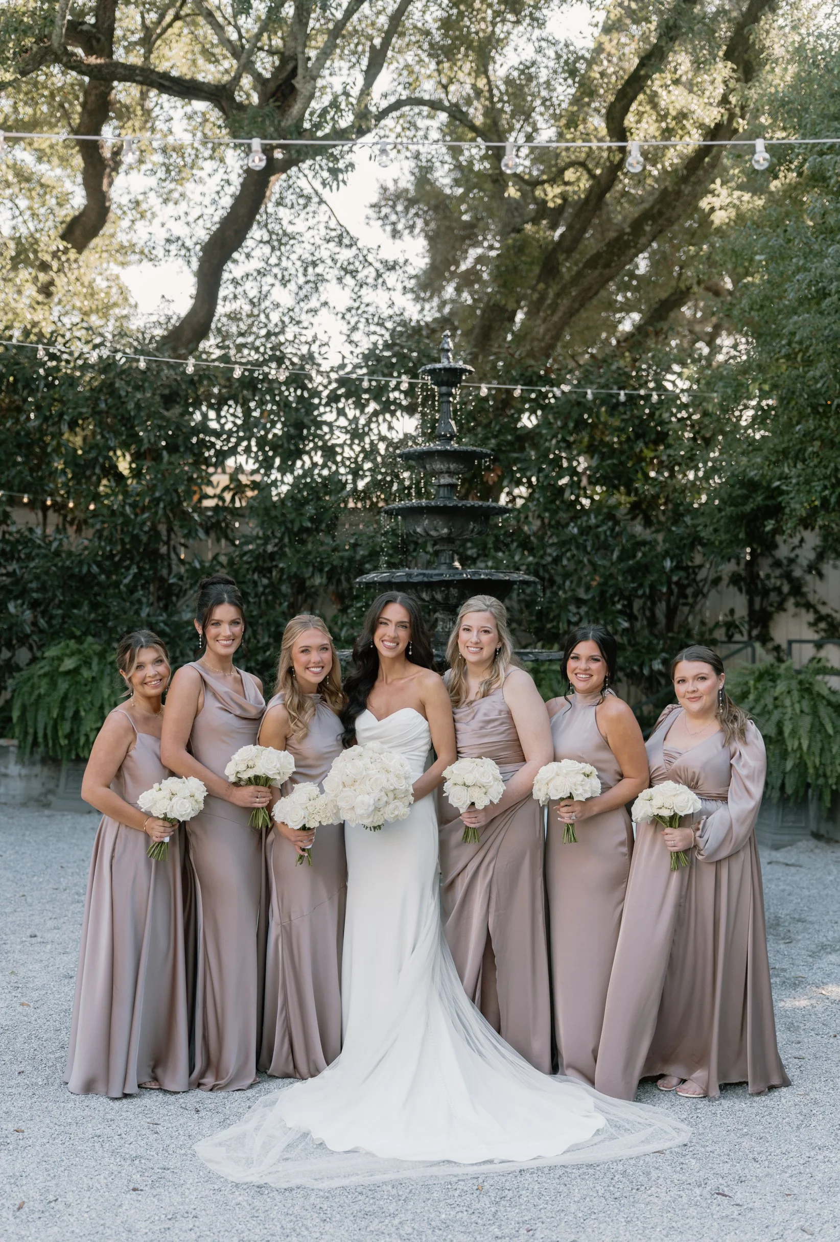 A bride in a white wedding gown surrounded by seven bridesmaids in matching taupe dresses, holding bouquets of white flowers, standing outdoors in front of a fountain and trees during sunset at a Pensacola Wedding Venue.