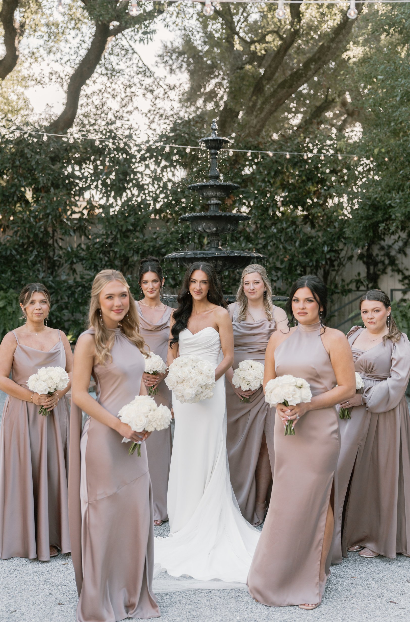 Bridal party of seven women and a bride posing outdoors in front of a fountain, with trees and string lights overhead, all dressed in elegant gowns and holding white bouquets.