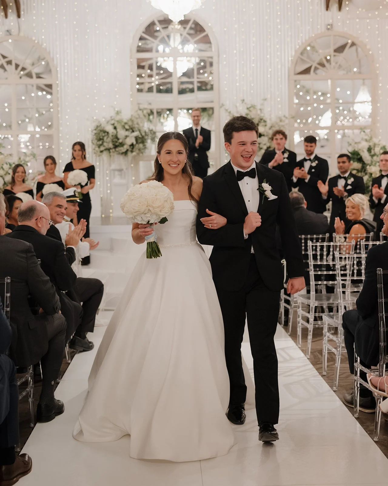 A bride and groom walking down the aisle at their wedding ceremony, smiling, with guests clapping and a wedding party behind them, decorated with flowers and lights.