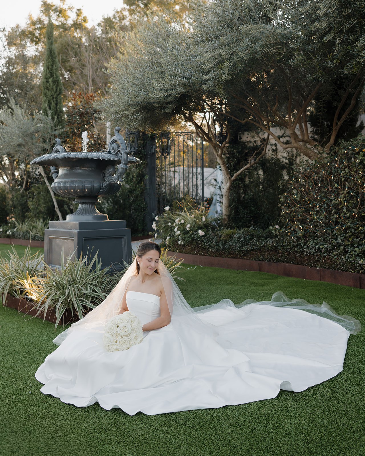 A bride in a white wedding gown sitting on green grass, holding a bouquet of white roses, outdoors in a garden with trees, bushes, and a large decorative fountain in the background.