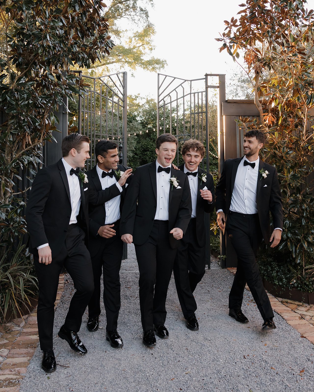 Group of five men in tuxedos walking and laughing together outdoors, with lush greenery and decorative wedding venue gate behind them.