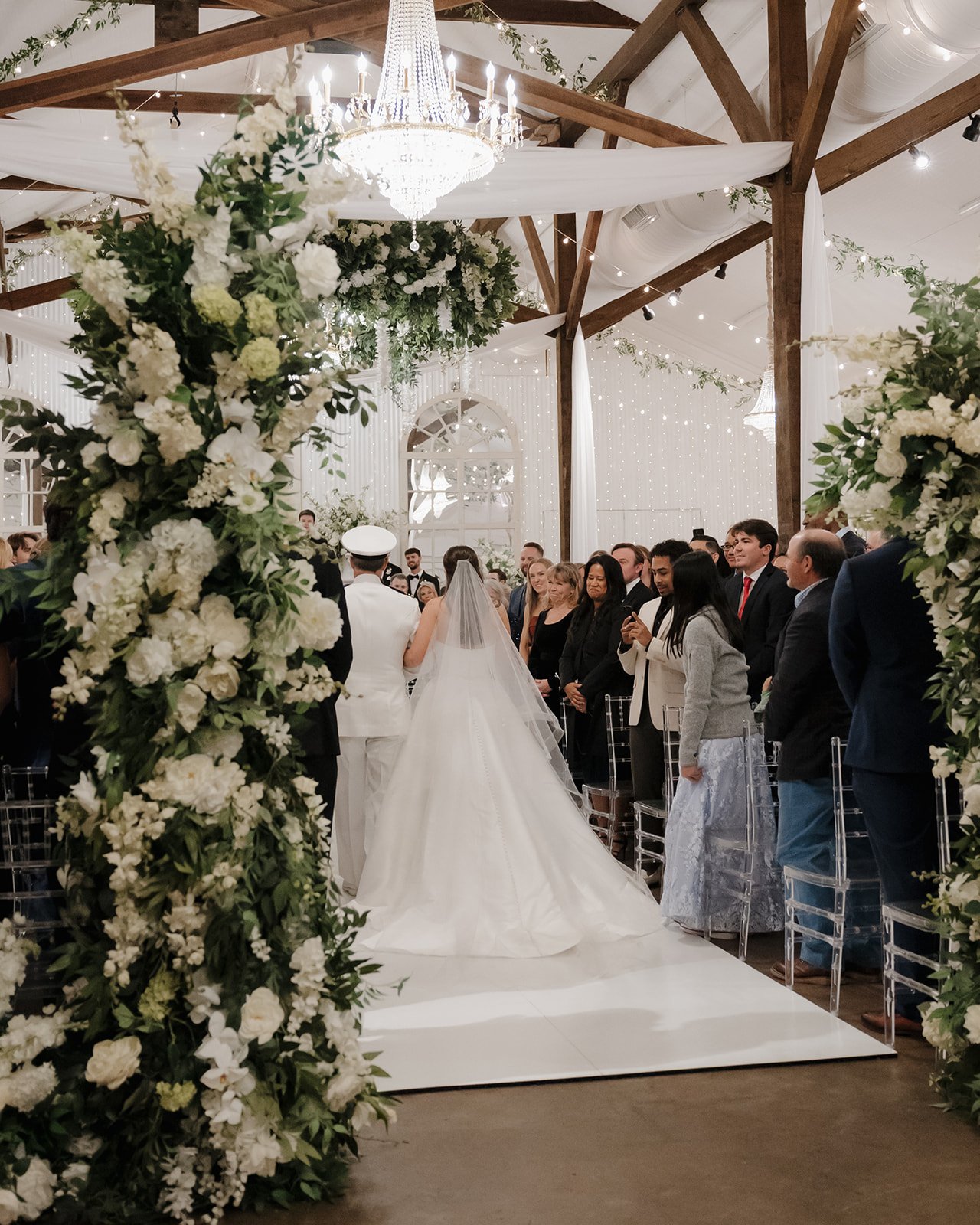 Wedding ceremony with bride in white gown and veil, groom in white military uniform, guests standing, decorated with white flowers and greenery, chandeliers and string lights hanging from wooden beams overhead, indoor Pensacola Wedding Venue