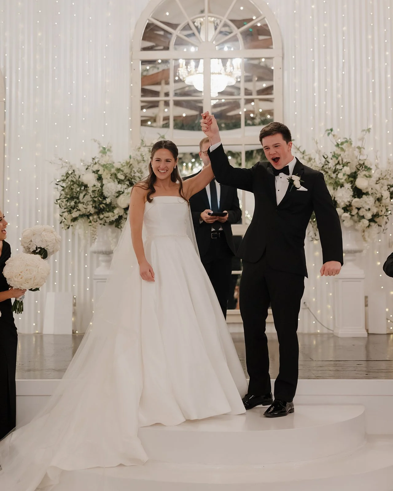 A newlywed couple at their wedding reception, raising their clasped hands in celebration. The bride is wearing a white wedding gown, and the groom is in a black tuxedo. They are smiling, with floral arrangements and fairy lights in the background.