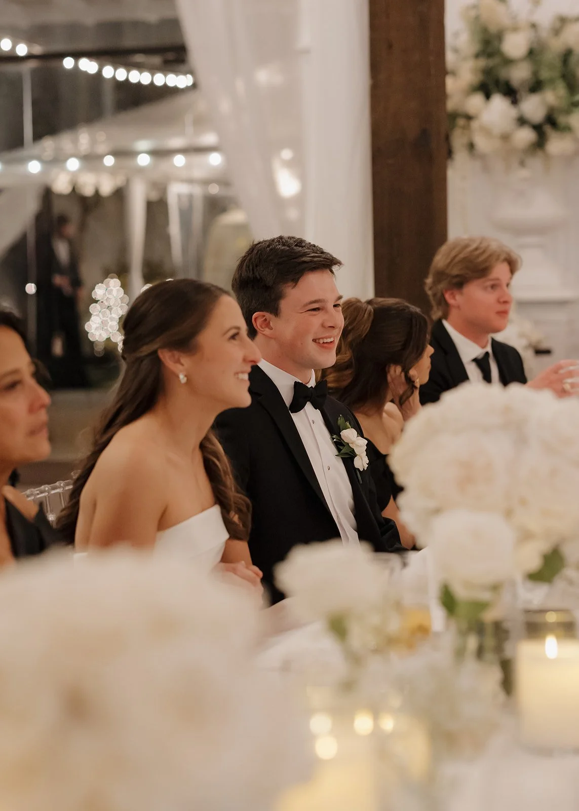 A group of people dressed in formal attire sitting at a wedding reception table, with the groom smiling and wearing a black tuxedo and boutonnière, and the bride in a strapless white dress.