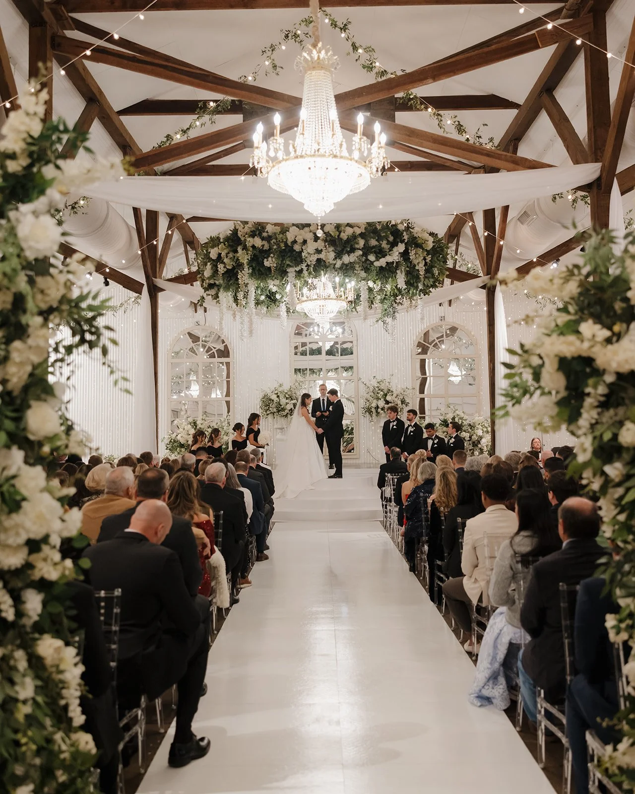 A Pensacola wedding ceremony taking place in an elegant, decorated hall with chandeliers, floral arrangements, and an audience watching two people exchanging vows at the altar.