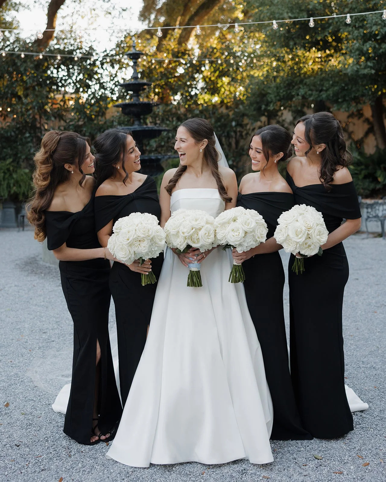 Bridal party at a wedding outdoors, with the bride holding a bouquet of white roses and her four bridesmaids, all wearing black off-the-shoulder dresses, standing on a gravel surface.