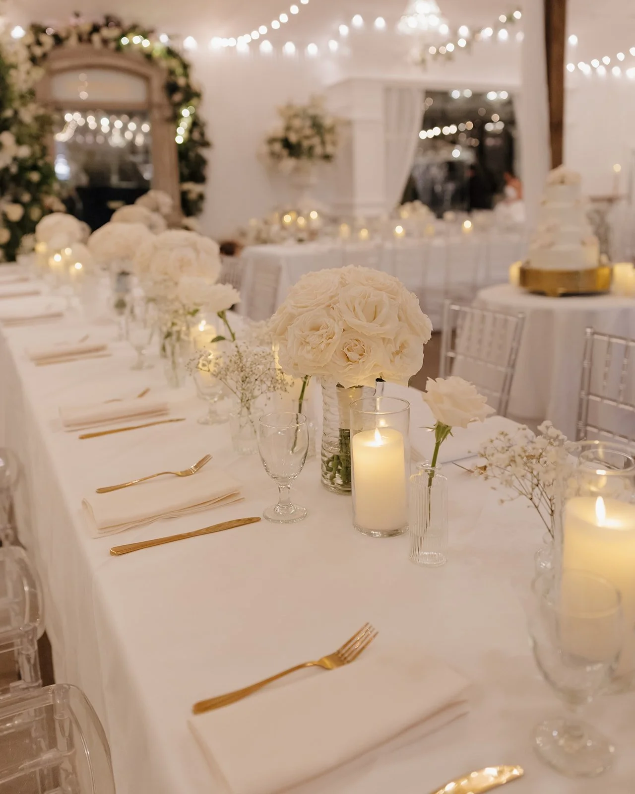 Elegant wedding reception table decorated with white flowers, candles, and gold cutlery in a softly lit room.