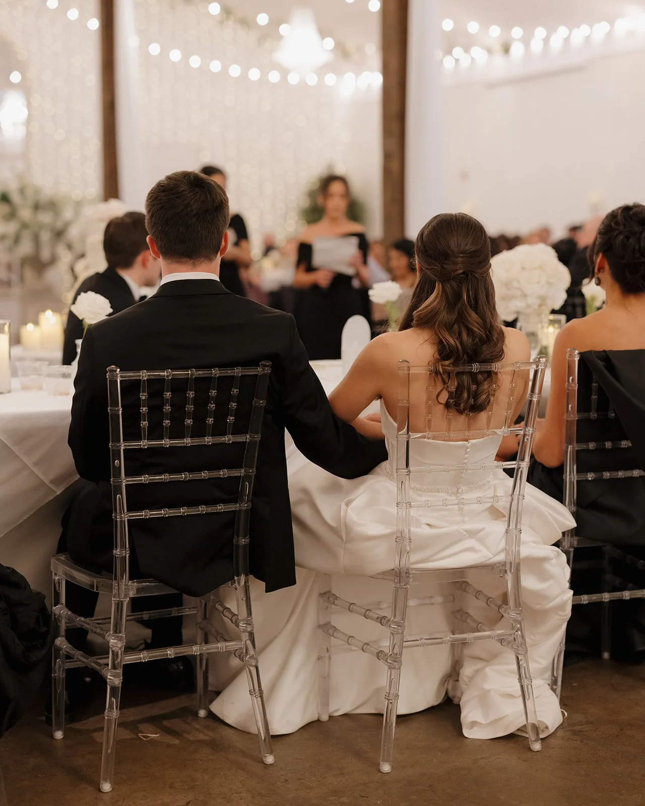 A bride and groom holding hands during their wedding reception, seated at a decorated head table surrounded by guests, with a woman giving a speech or reading in the background.