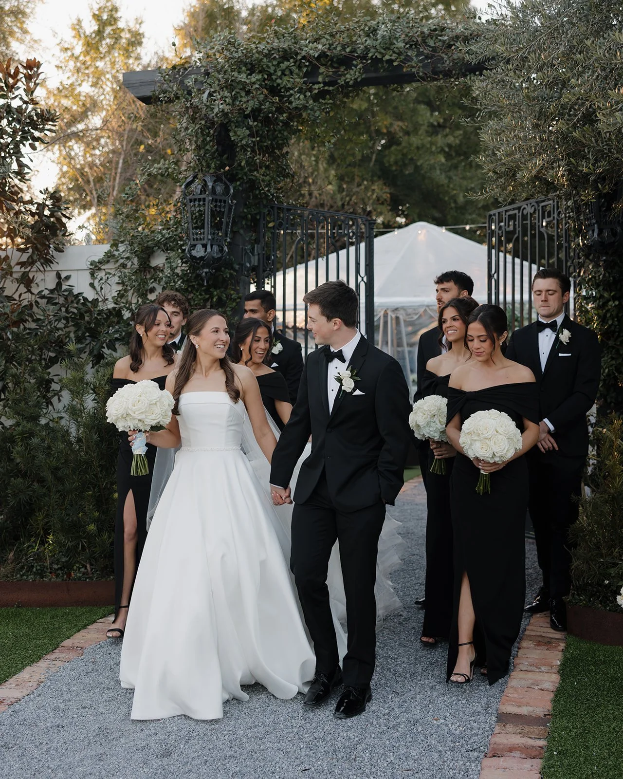 A wedding party walking under a black gate with greenery, with the bride and groom holding hands and smiling at each other, surrounded by bridesmaids and groomsmen in formal attire, outdoors during daytime.