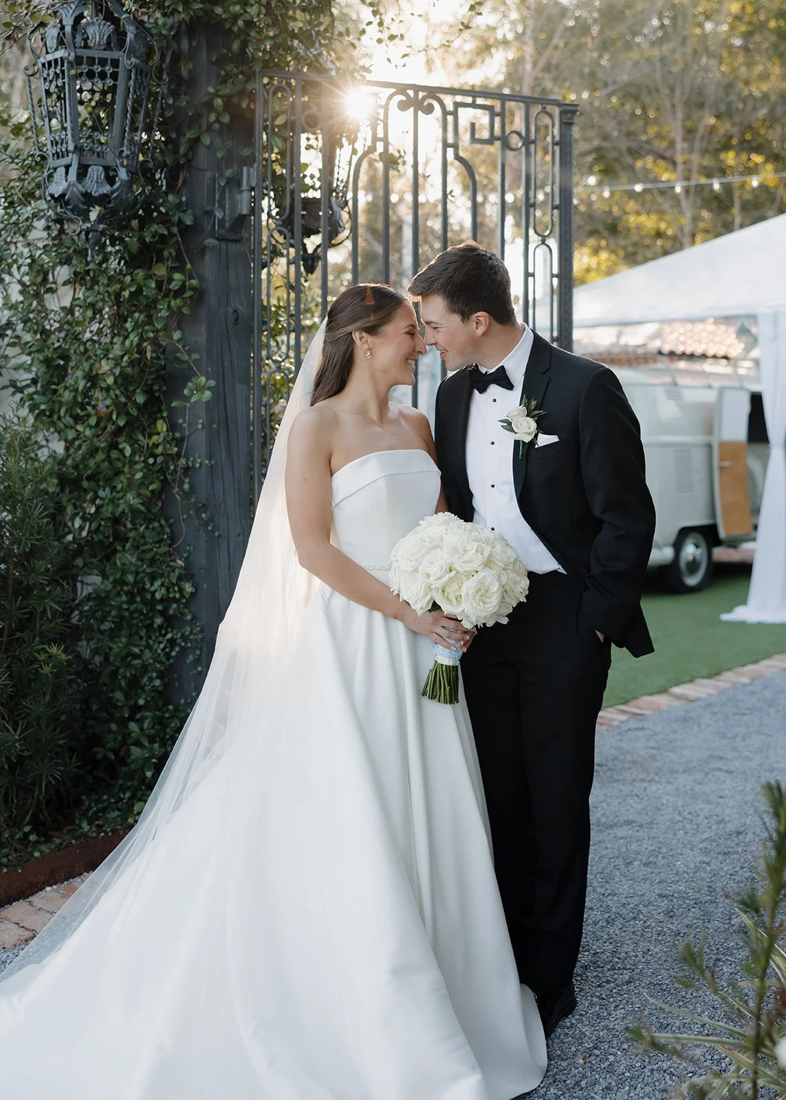 A bride and groom are standing close together outdoors, smiling with their foreheads touching. The bride is wearing a strapless white wedding gown and holding a bouquet of white roses, while the groom is in a black tuxedo with a bow tie and a white b