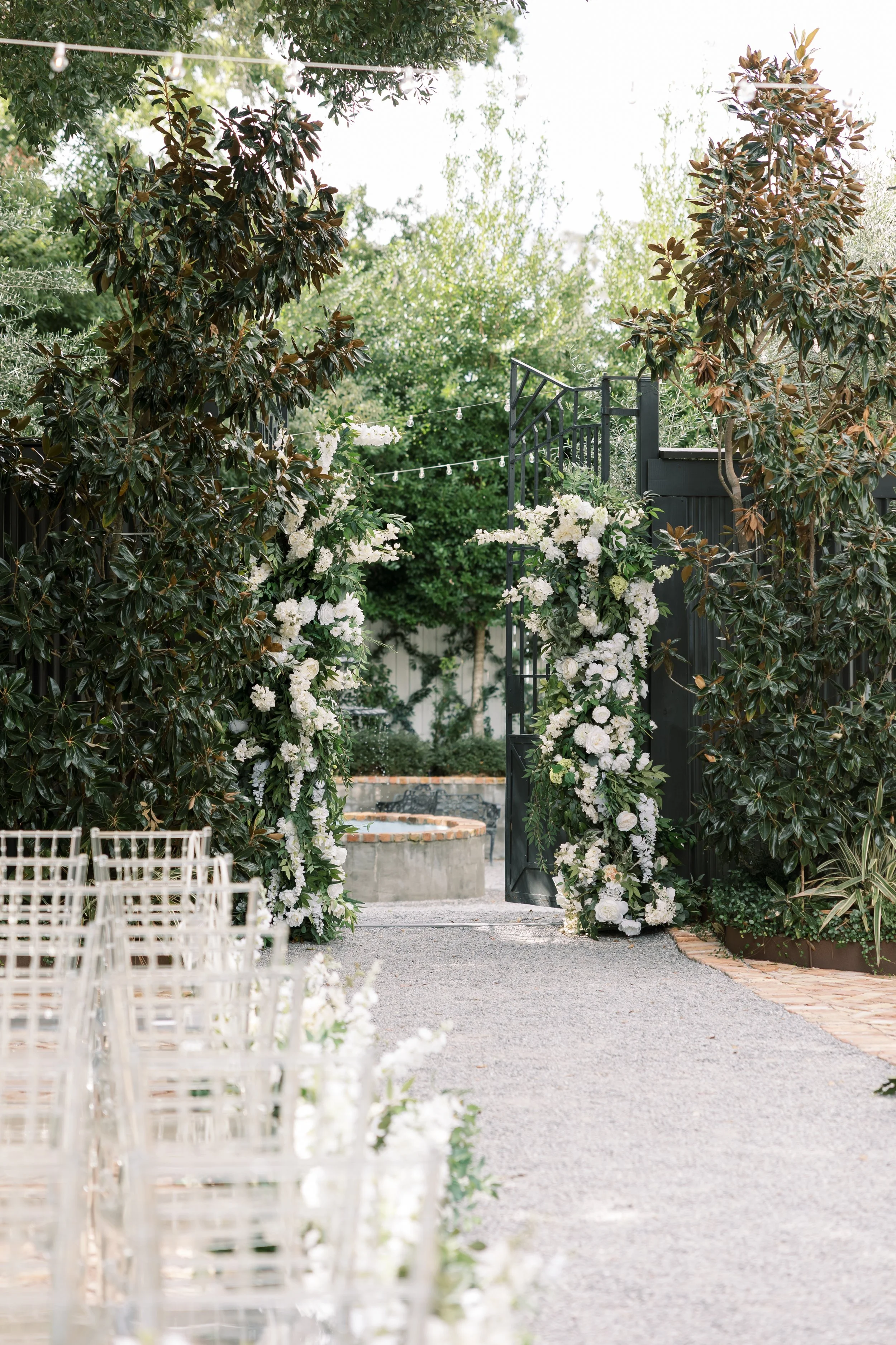Decorated outdoor wedding arch with white flowers, pathway leading to the arch, and chairs on the side, set in a lush green garden.
