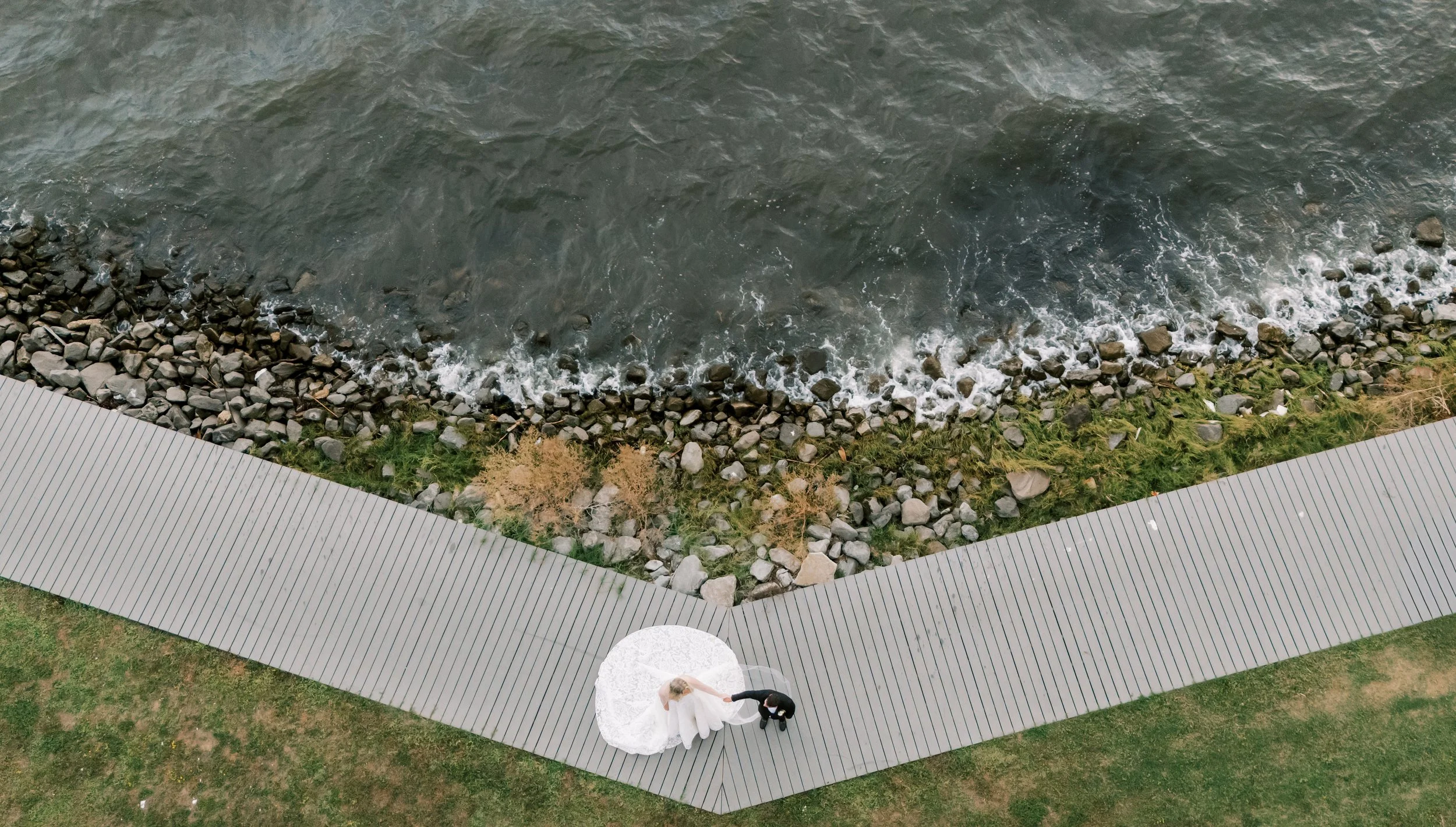 A bride in a white wedding dress and a groom in a black suit holding hands under a white umbrella on a gray wooden walkway next to a rocky waterfront.