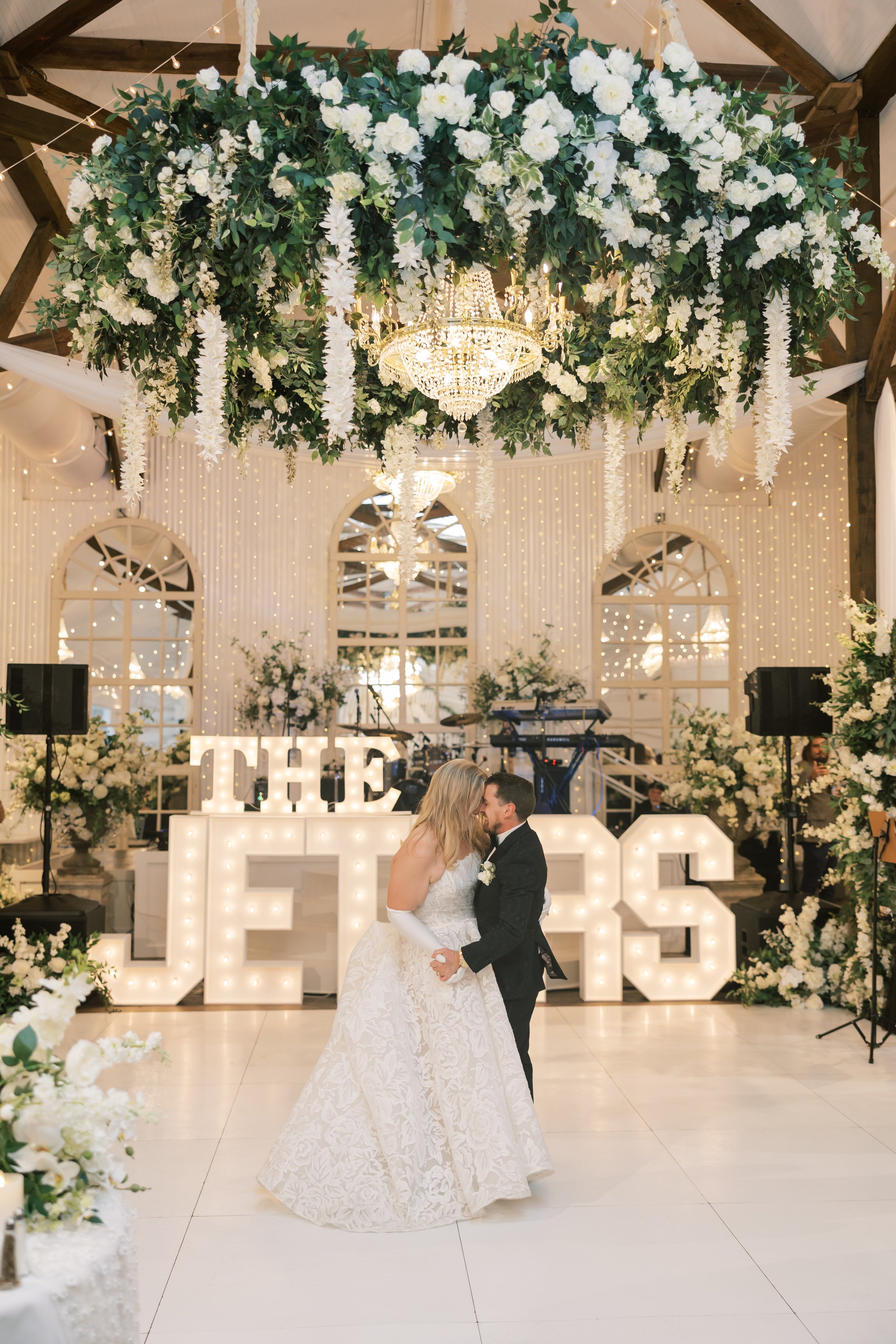 A bride and groom sharing a dance at their wedding reception, decorated with white flowers and illuminated letters in the background.
