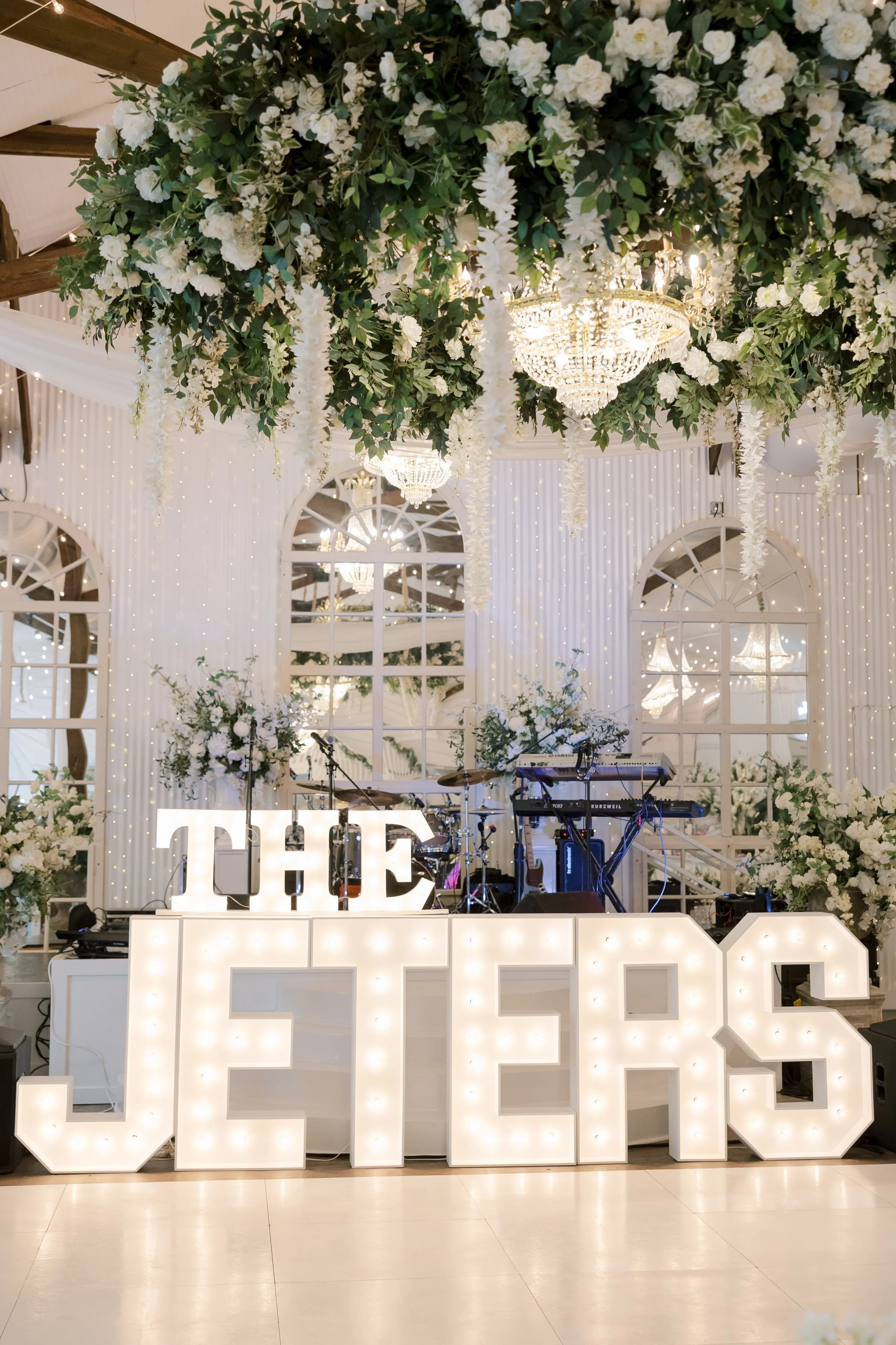 Elegant wedding reception with large illuminated signs reading 'THE' and 'JEN'S' in front of a stage with musical instruments, surrounded by white floral arrangements and hanging greenery, with crystal chandeliers and fairy lights in the background.